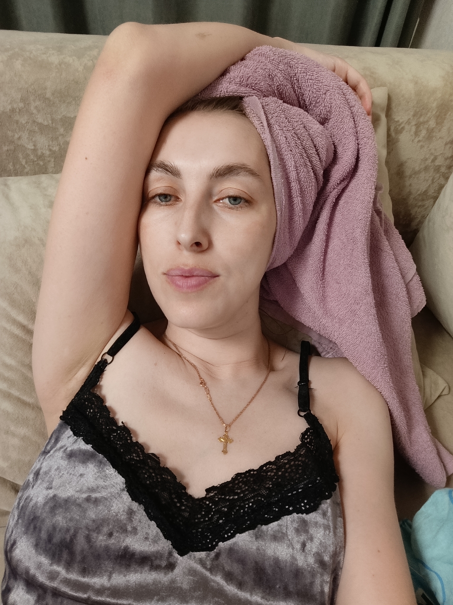 A woman is lying on a beige couch, wearing a gray velvet top with black lace trim and a gold necklace with a cross pendant. Her hair is wrapped in a pink towel. She is fair-skinned, with blue eyes, and appears to be looking directly at the camera. The background includes a gray curtain. The location is Chaikovsky, Russia.