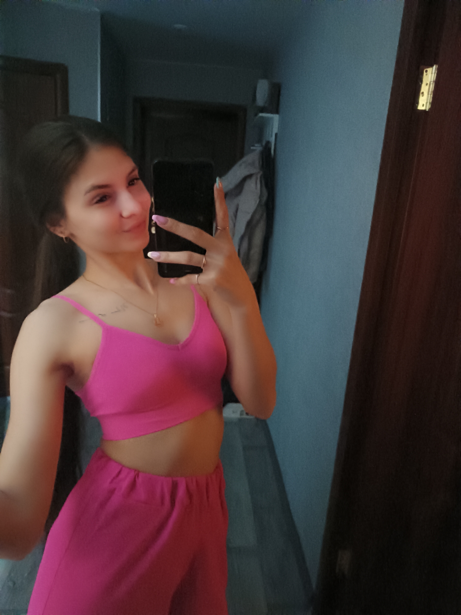 A young woman is taking a selfie in what appears to be an indoor setting, possibly a bedroom or hallway. She is wearing a matching pink crop top and pants. She is holding a smartphone in her right hand, obscuring part of her face. The background includes a blue wall, a door with a noticeable hinge, and a glimpse of another room with hanging clothing. The location is unknown.