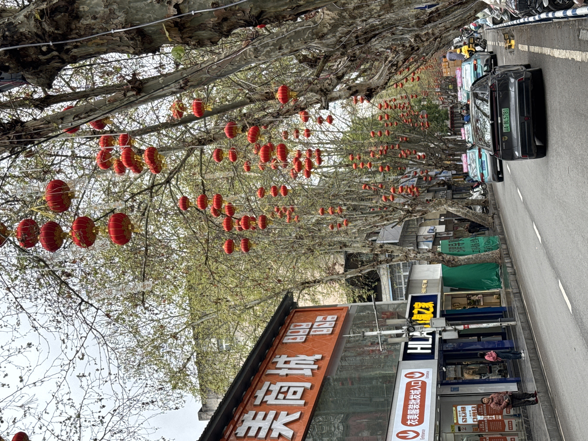 The image depicts a street in Qianjiang, China, decorated with red lanterns hanging from trees lining the roadside. On the right, cars are parked along the street, and people are walking on the sidewalk in front of stores. Storefront signs are visible, including one that reads "Mei Shang Cheng," indicating the name of a merchant shop.