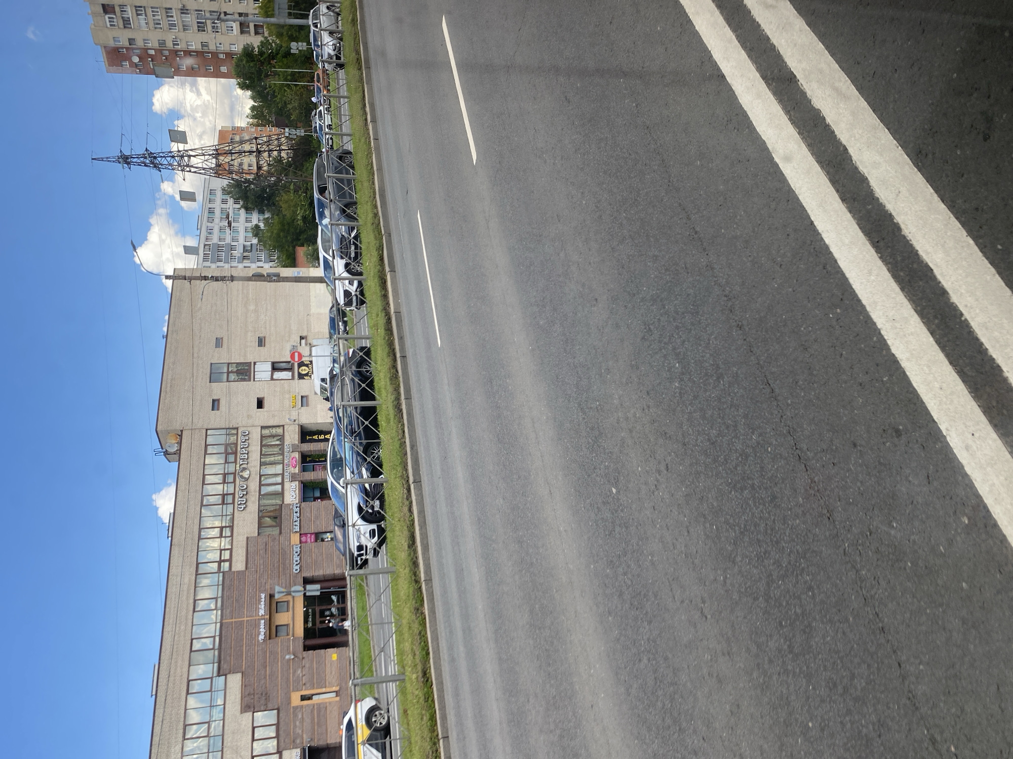 A bright, sunny day in Saint Petersburg, Russia, is depicted in this image. The photo captures a street scene with cars parked along the side of a road. A modern building with large windows and a patterned facade dominates the background. Signs on the building indicate various businesses, including "Фото Твердито" (Foto Tverdi to), "ЦВЕТЫ" (Flowers), "ОРТОПЕДИЯ" (Orthopedics), and "МАРИЯ" (Maria). Several cars are parked in front of the building, with a fence separating the parking area from the sidewalk. The road in the foreground has clear white lane markings. In the distance, a tall electrical tower and apartment buildings are visible, suggesting an urban environment. The sky is blue with scattered white clouds.