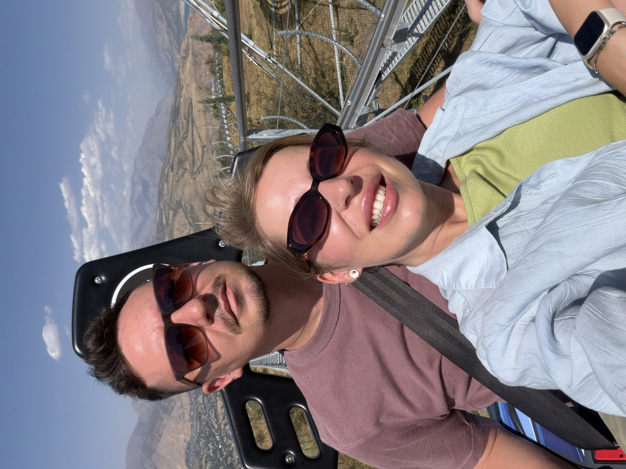 A man and a woman are seated side-by-side in a black, multi-person cart, likely on a mountain coaster, with a scenic mountainous landscape in the background. The man, wearing sunglasses and a maroon t-shirt, is positioned behind and to the left of the woman. The woman, also wearing sunglasses and a light blue collared shirt over a green top, is in the foreground, smiling at the camera. Both appear to be wearing seatbelts. The cart is on a track, which is visible curving down the mountainside. The environment is dry and arid, with sparse vegetation and a clear blue sky. The image was taken in Charvak, Uzbekistan.