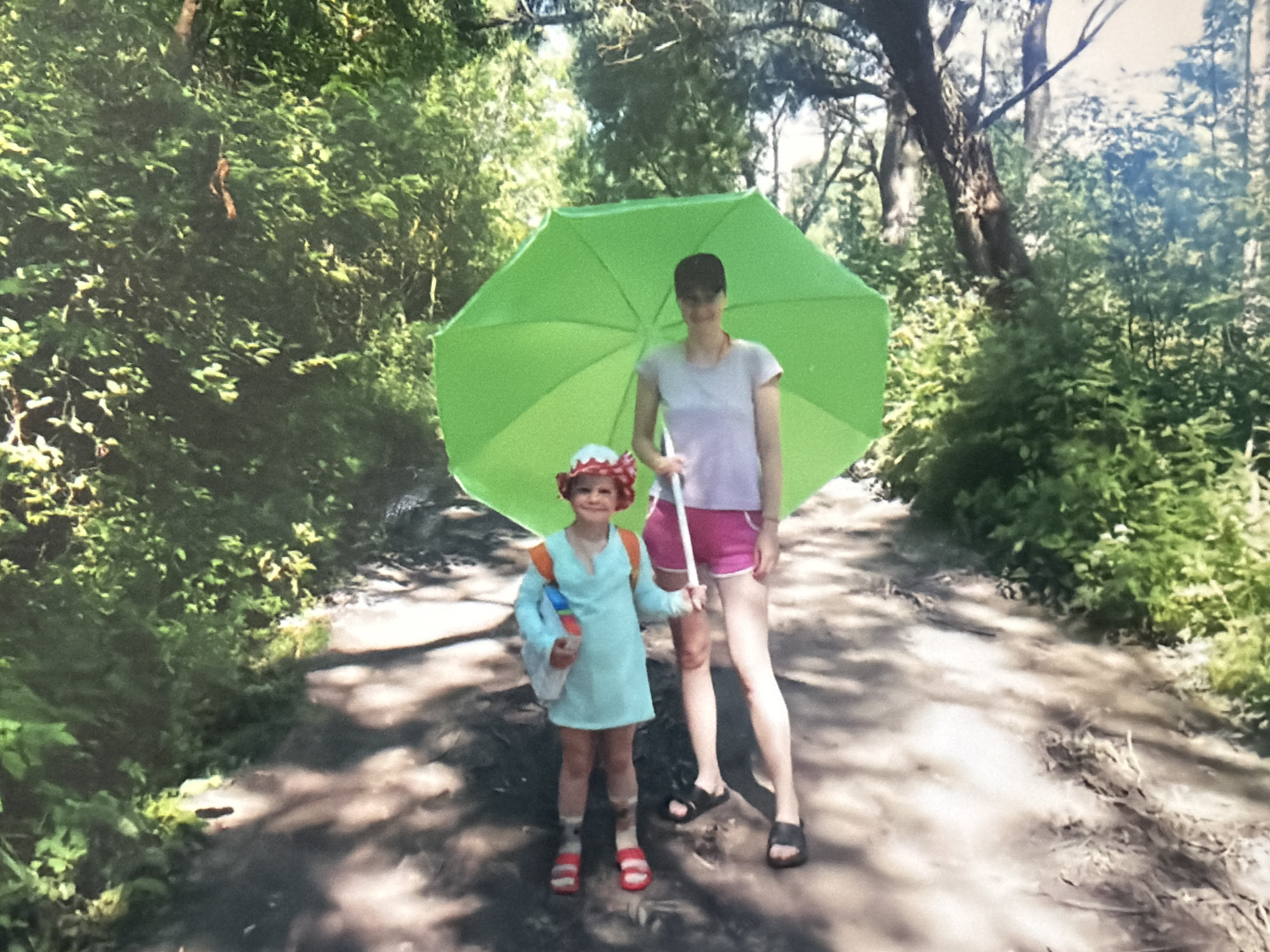 A photograph shows a child and an adult standing on a dirt path through a forest. The adult, likely a woman, is holding a green umbrella over both of them. The child is wearing a light blue dress-like garment, a red and white hat, and red sandals. The adult is wearing a light gray t-shirt, pink shorts, and black sandals. The forest environment includes various trees and shrubs. The location is likely a park or nature trail.