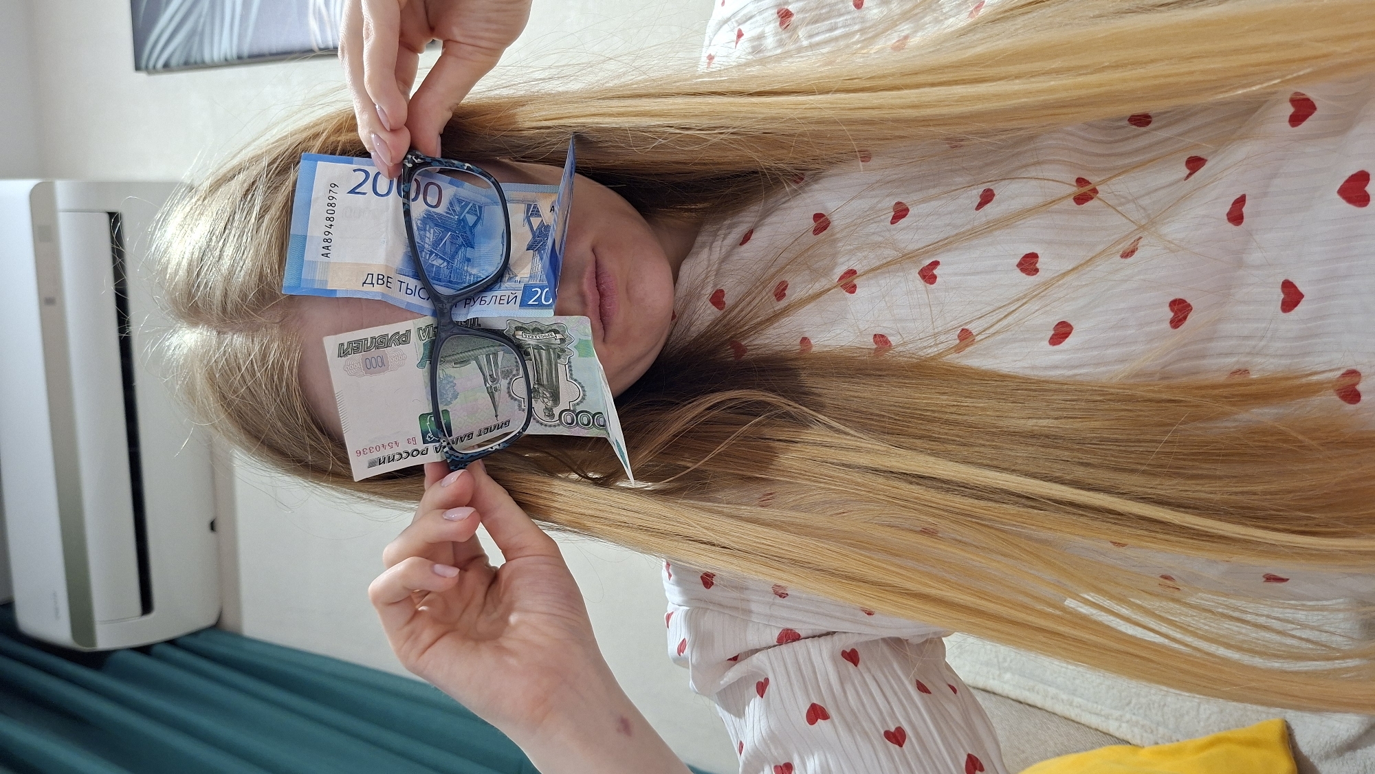 Here's a detailed description of the image:
**People:** The image features a person with long, blonde hair. Only parts of their face (eyes covered by money, lips) are visible. Two hands are present, holding up eyeglasses and two Russian Ruble banknotes. The person is wearing a white top or shirt with a pattern of small red hearts.
**Subjects:** The main subjects are the person, the Russian money (Rubles), and the glasses.
**Objects:** The visible objects include:
* Two Russian Ruble banknotes: one 2000 Ruble note (blue) and one 1000 Ruble note (green)
* A pair of eyeglasses with a patterned frame.
* The person's hair and clothing.
* A white, box-shaped object (likely an air purifier or similar appliance) in the background.
* A section of a blue-green curtain or bedspread.
* A yellow pillow.
* A painting on the wall
**Actions:** The hands are holding the banknotes and glasses in front of the person's face, partially obscuring their eyes.
**Interactions:** The interaction appears to be a playful pose, with the money and glasses used as humorous props.
**Environment:** The setting appears to be indoors, likely a bedroom or living area, given the clothing, curtain, and home appliances visible.
**OCR information:**
The OCR in the image shows information printed on the banknotes, including the denominations ("2000" and "1000"), currency name ("РУБЛЕЙ"- Rubles), and serial numbers (e.g., "AA894808979", "Б3 4540336").
**Location:** There is no information suggesting this image comes from the city of Chaikovsky, Russia.