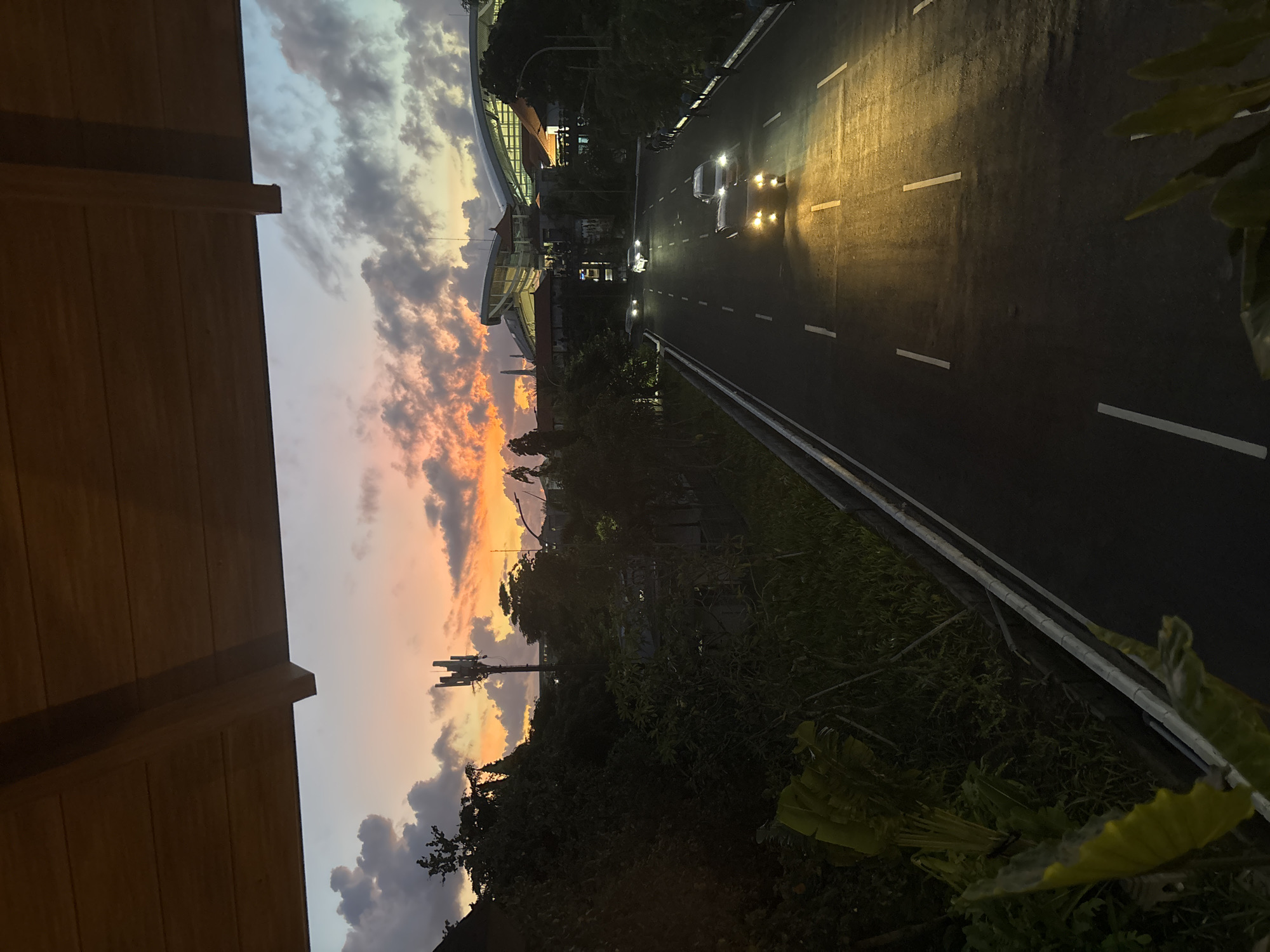 The image depicts a dusk scene overlooking a road in Kuta, Indonesia. The sky is a mix of colors, from blue and gray to orange and pink, reflecting the setting sun behind a cloud cover. A building with architectural details is visible, positioned adjacent to the road, which has several cars with headlights. Vegetation lines the lower portion of the image, obscuring some details of the scene. The road is a dark paved surface with white lane markings. A wooden structure partially frames the view from the left side. A cell tower is located in the middle-ground.