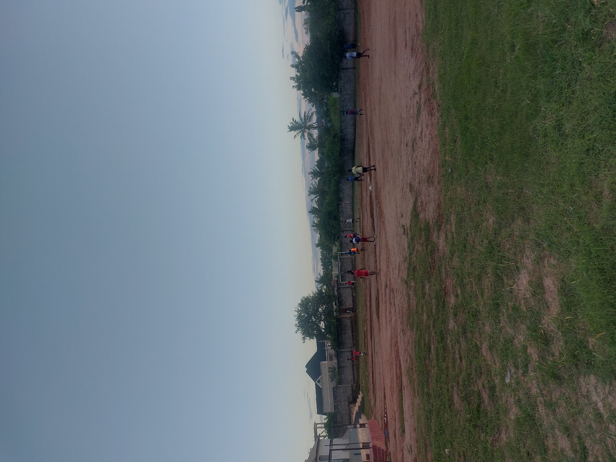 The image depicts a dusty field in Dar es Salaam, Tanzania, next to a patch of green grass. Several individuals, possibly children, are engaged in an activity on the field, perhaps playing soccer. A low wall borders the field on one side, with buildings visible beyond it. In the distance, a line of palm trees and other vegetation is present. The sky is a pale blue, suggesting either early morning or late afternoon.