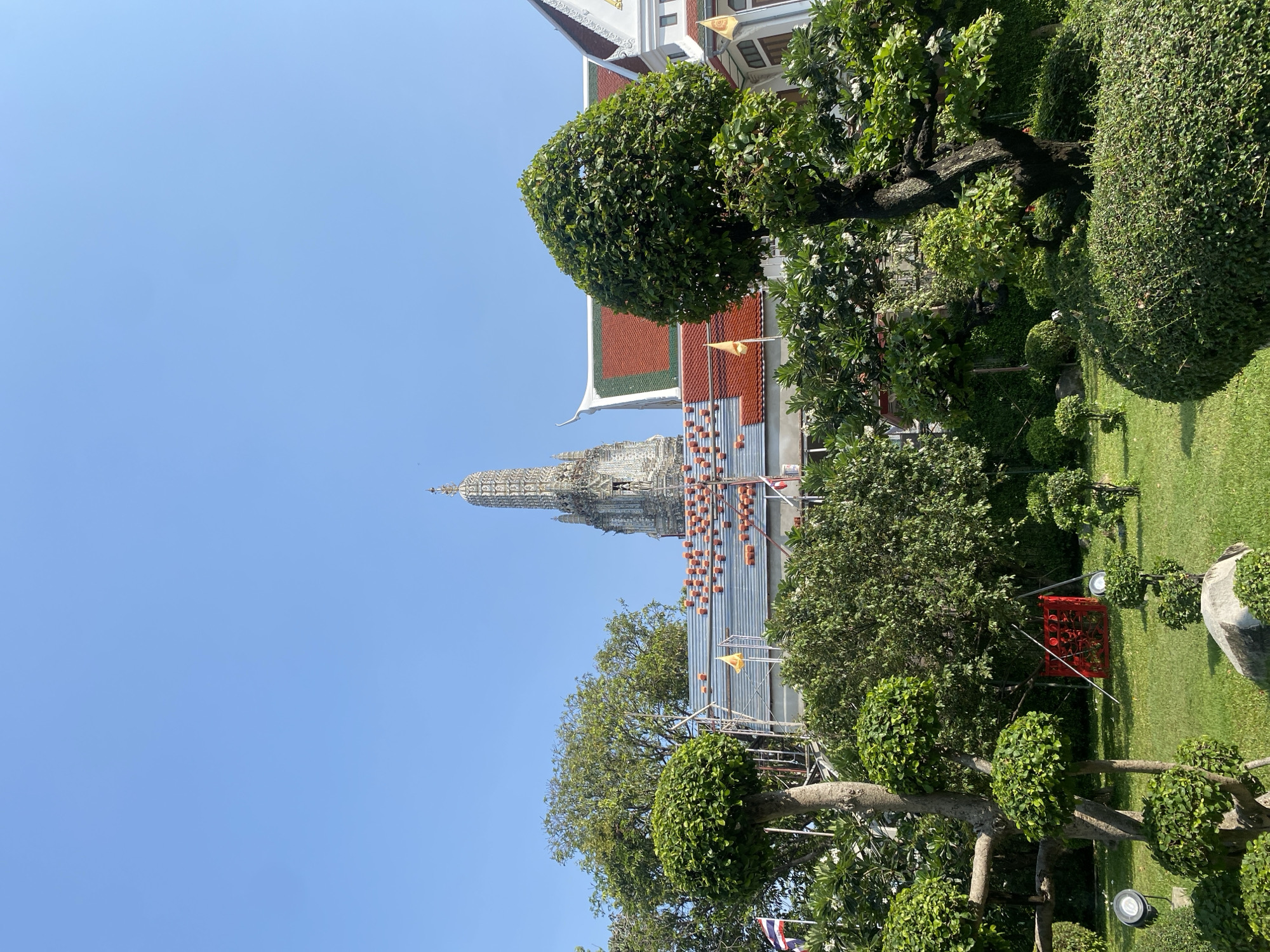 The image depicts Wat Arun Ratchawararam Ratchawaramahawihan (Temple of Dawn) in Bangkok, Thailand. The tall, white, ornate pagoda is partially obscured by lush green trees and manicured shrubbery. A red-tiled roof of an adjacent building is visible on the right side of the frame. The sky is clear and blue. A temporary scaffolding and some materials are visible, indicating ongoing maintenance or construction.