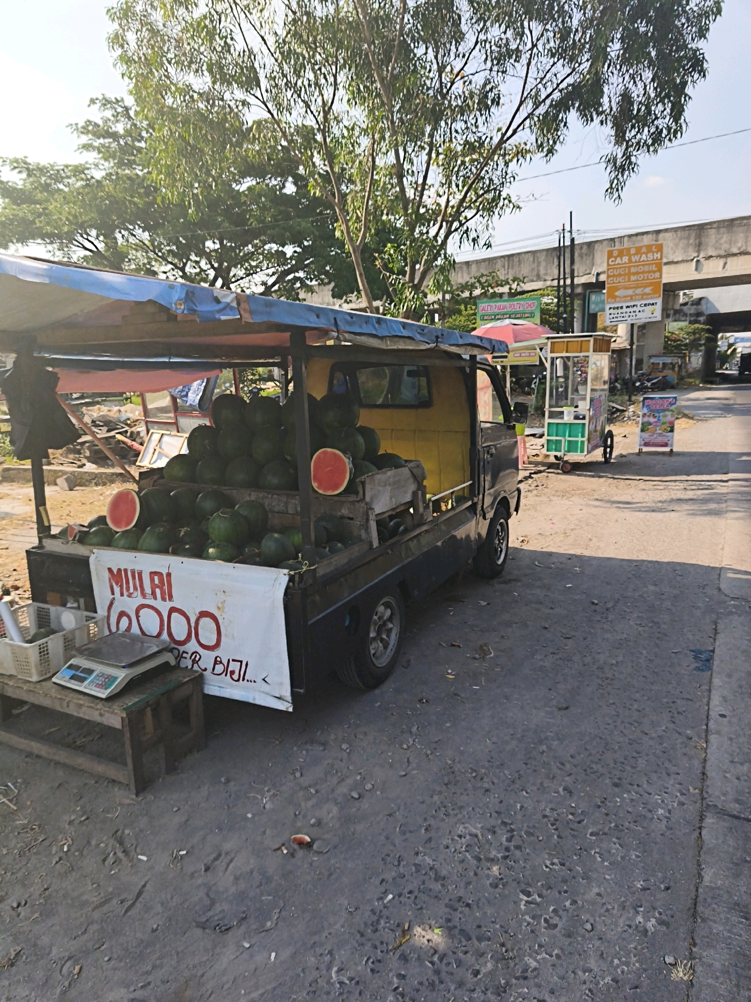 A street vendor in Desa Dibal, Indonesia, is selling watermelons from a small truck. The truck has a yellow cab and a dark-colored bed covered with a blue tarp. A large quantity of whole watermelons is piled in the truck bed, with two halved watermelons displayed on top. A white banner is affixed to the front of the truck bed, displaying text that appears to be pricing information, with "MULAI" (meaning "start") and "6000" prominently visible. Beside the truck, a small wooden stand holds a white plastic basket and a digital scale. In the background, an elevated road structure is visible, along with other small vendor stalls and signage, including one for "CAR WASH CUCI MOBIL CUCI MOTOR." The ground is paved with asphalt and shows signs of wear and scattered debris. Trees with green foliage line the background. The scene is illuminated by natural sunlight, casting shadows on the ground.