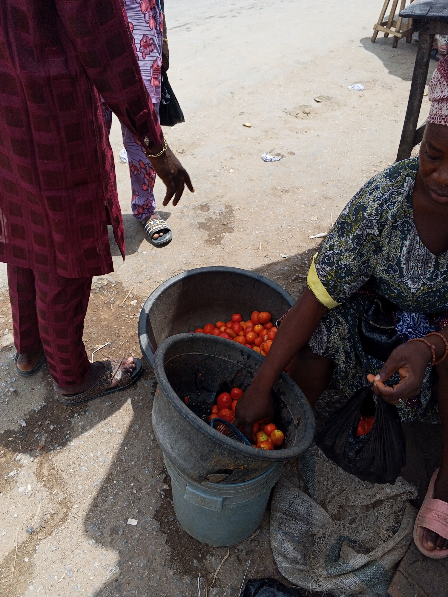 In Zango-ndaji, Nigeria, a street scene depicts a woman selling tomatoes. She is crouching beside a stack of three buckets, the top two filled with red tomatoes. She is filling a black plastic bag with tomatoes. A customer stands beside her, their hand reaching out towards the tomatoes. Another person stands nearby. The scene takes place on a dirt street with some trash scattered around.
