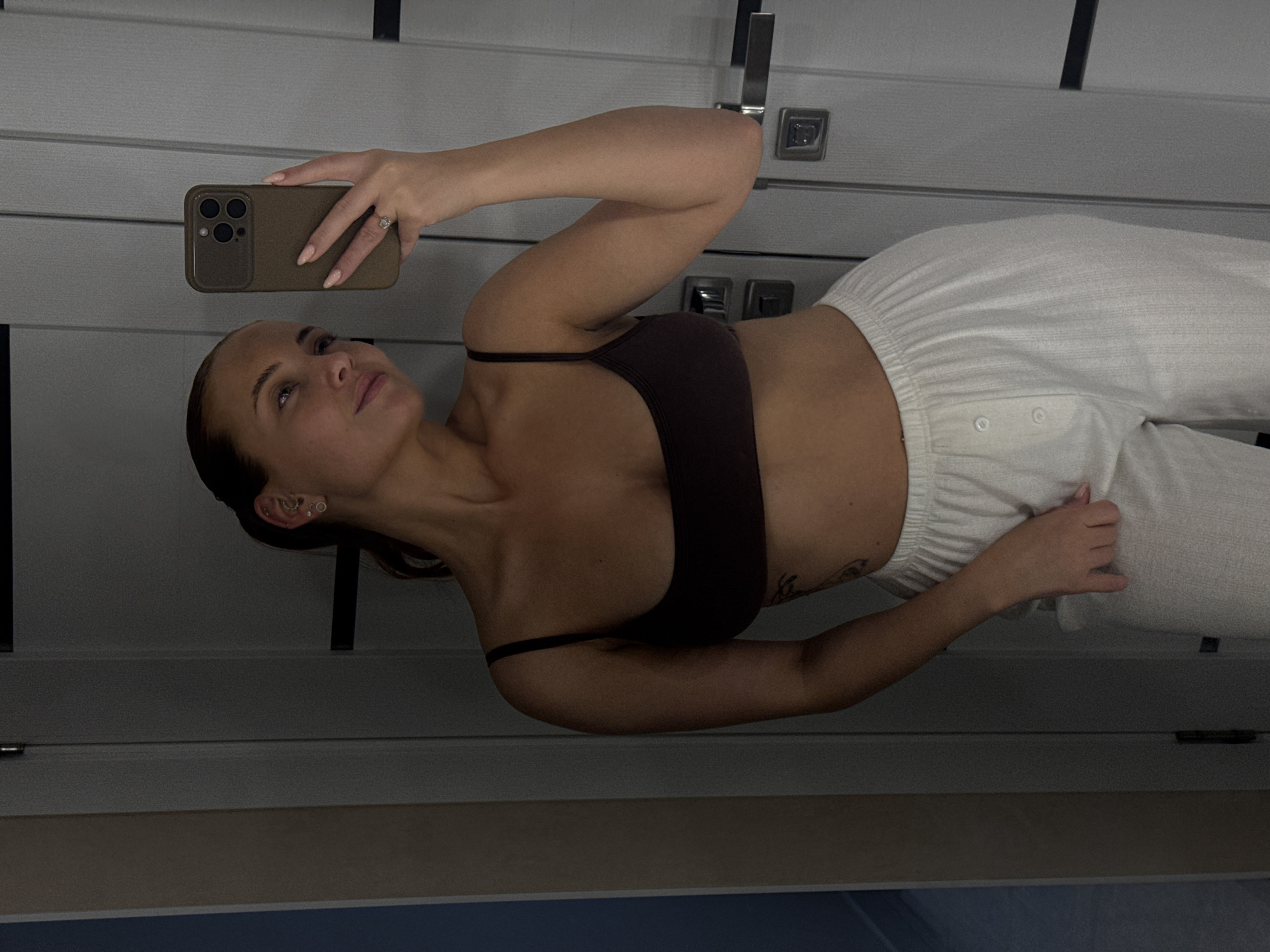 This image, taken in Chaikovsky, Russia, portrays a young woman taking a self-portrait indoors. She is positioned to the left side of the frame, holding a smartphone with a brown case in her right hand; a ring is visible on her ring finger. Her left hand rests casually on her hip.
The woman has tanned skin and her dark hair is pulled back. She is dressed in a dark brown or black bralette or crop top paired with light-colored, possibly white, wide-leg pants that feature decorative buttons near the waist. Multiple piercings adorn her left ear, and a faint tattoo can be seen on her left arm. Her expression is relaxed and neutral, with her gaze directed slightly upwards and to the side.
The background consists of a light gray or white paneled wall or door, equipped with dark, modern door handles. A dark horizontal element runs across the upper part of the background, and a narrow, lighter strip, likely part of the floor or skirting, is visible at the bottom. The lighting is soft and ambient, characteristic of an indoor setting. No visible text is present in the image.