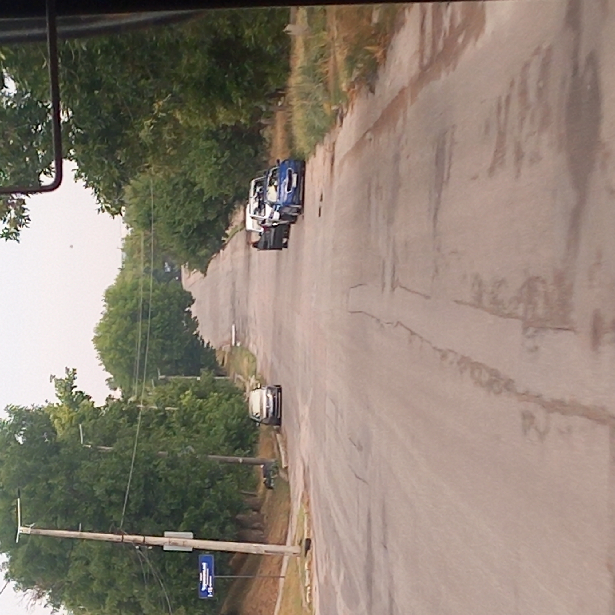 The image captures a view of a paved road in Kropyvnytskyi, Ukraine, lined with trees on both sides. The road appears to be in a residential area, with a few cars parked along the side. In the foreground, the road surface shows signs of wear and tear with cracks and some darker patches, possibly indicating water or oil stains.
On the left side of the road, a utilitarian pole with wires and a blue street sign is visible. The sign has white lettering and appears to indicate street names or directions. Further down the road on the left, a dark-colored sedan is parked.
On the right side of the road, a group of vehicles is parked. These include a white van or minivan, and at least two other cars, one of which is a blue convertible. The blue convertible is the most prominent vehicle on the right, parked facing away from the viewer. The area beside the road on the right is overgrown with tall grass and some scattered foliage.
The overall impression is of a quiet, tree-lined street with parked cars, suggesting a normal day in a residential neighborhood. The sky is overcast, and the lighting is diffused. The perspective seems to be from a vehicle, possibly a bus or a car, given the partial view of what appears to be a side mirror frame at the top right corner of the image.
