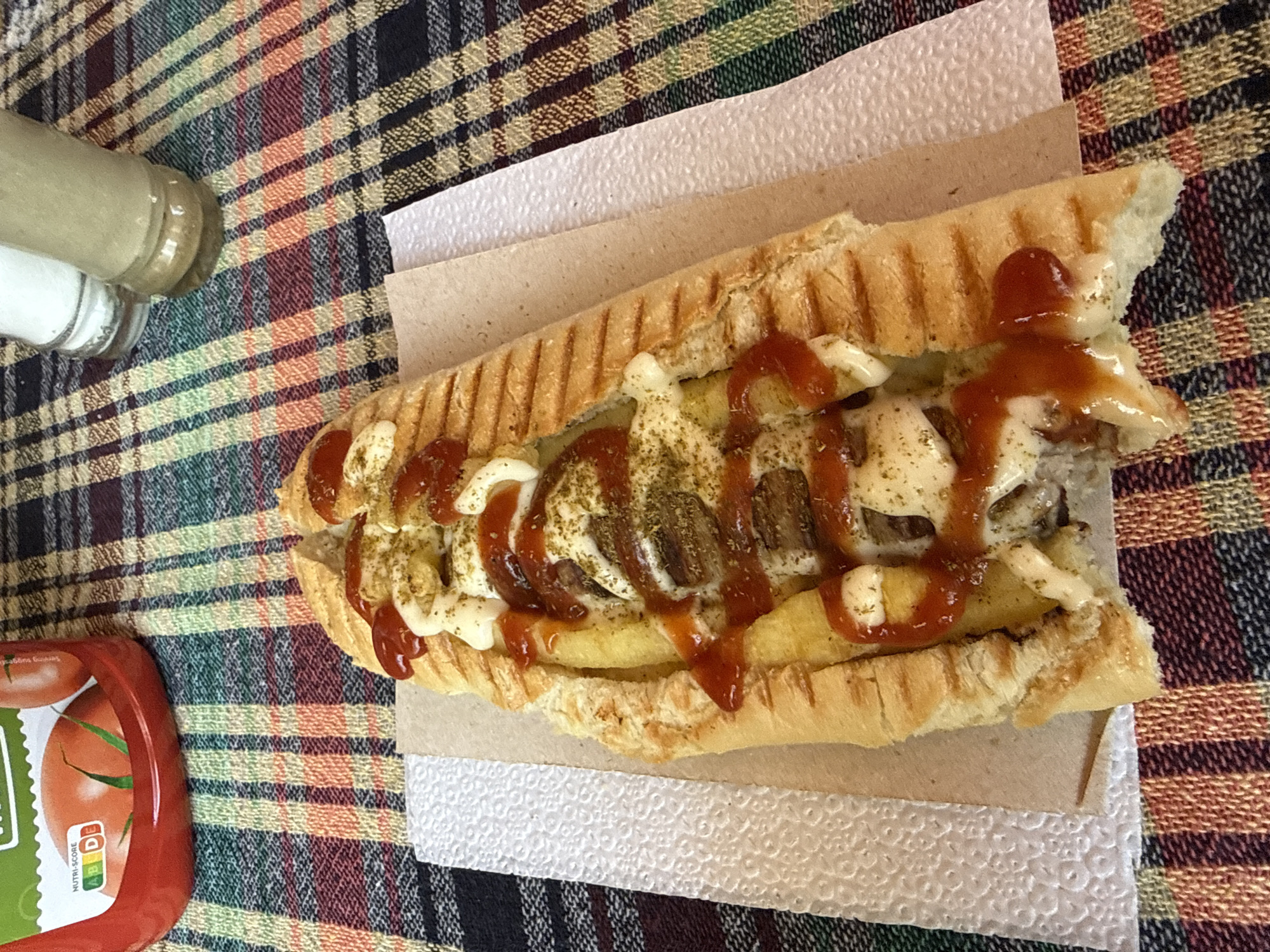 The image depicts a sandwich, possibly a panini, topped with ketchup, mayonnaise, and spices, resting on a multi-layered paper towel and cardboard. The setting is a table covered with a checkered tablecloth. In the periphery, there are condiment bottles and a ketchup bottle visible. There is no immediate indication of the specific location in Elhovo, Bulgaria, though the style of food preparation and presentation might suggest it's a local eatery. The scene is static with no visible human presence or activity.