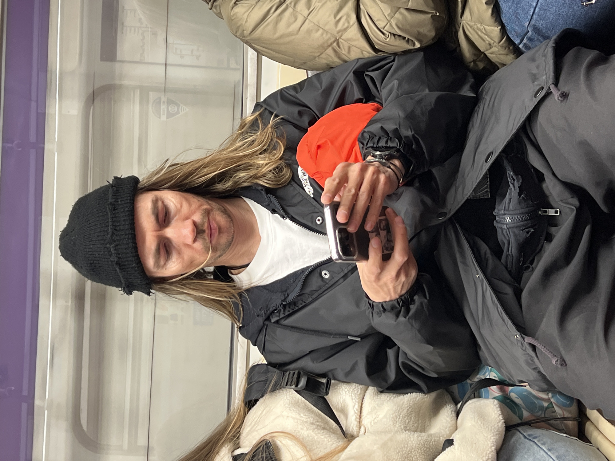 A person with long blond hair wearing a black beanie, white t-shirt, and black jacket is holding a smartphone. The person is seated, possibly on public transportation. An orange object is visible behind the person. The location is Saint Petersburg, Russia, discernible from the public transit interior. Other passengers and their belongings surround the person.