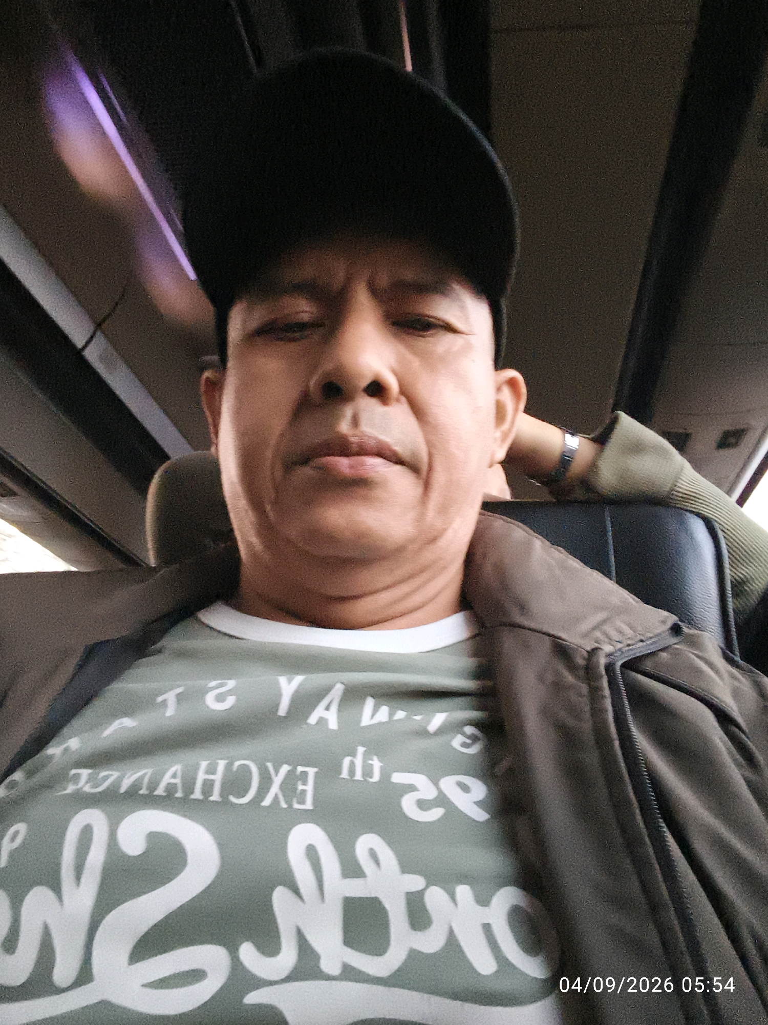 A man wearing a black baseball cap and a green t-shirt with white lettering sits in a vehicle. He is looking towards the camera, and his face is partially obscured by the cap and the angle of the photo. The background appears to be the interior of a bus or van, with seats visible. A date and time stamp in the bottom right corner reads "04/09/2026 05:54". The man has a wristband on his left arm. The t-shirt has text that appears to be "T G YA" and "DOYAMCHOXE" and "m2".