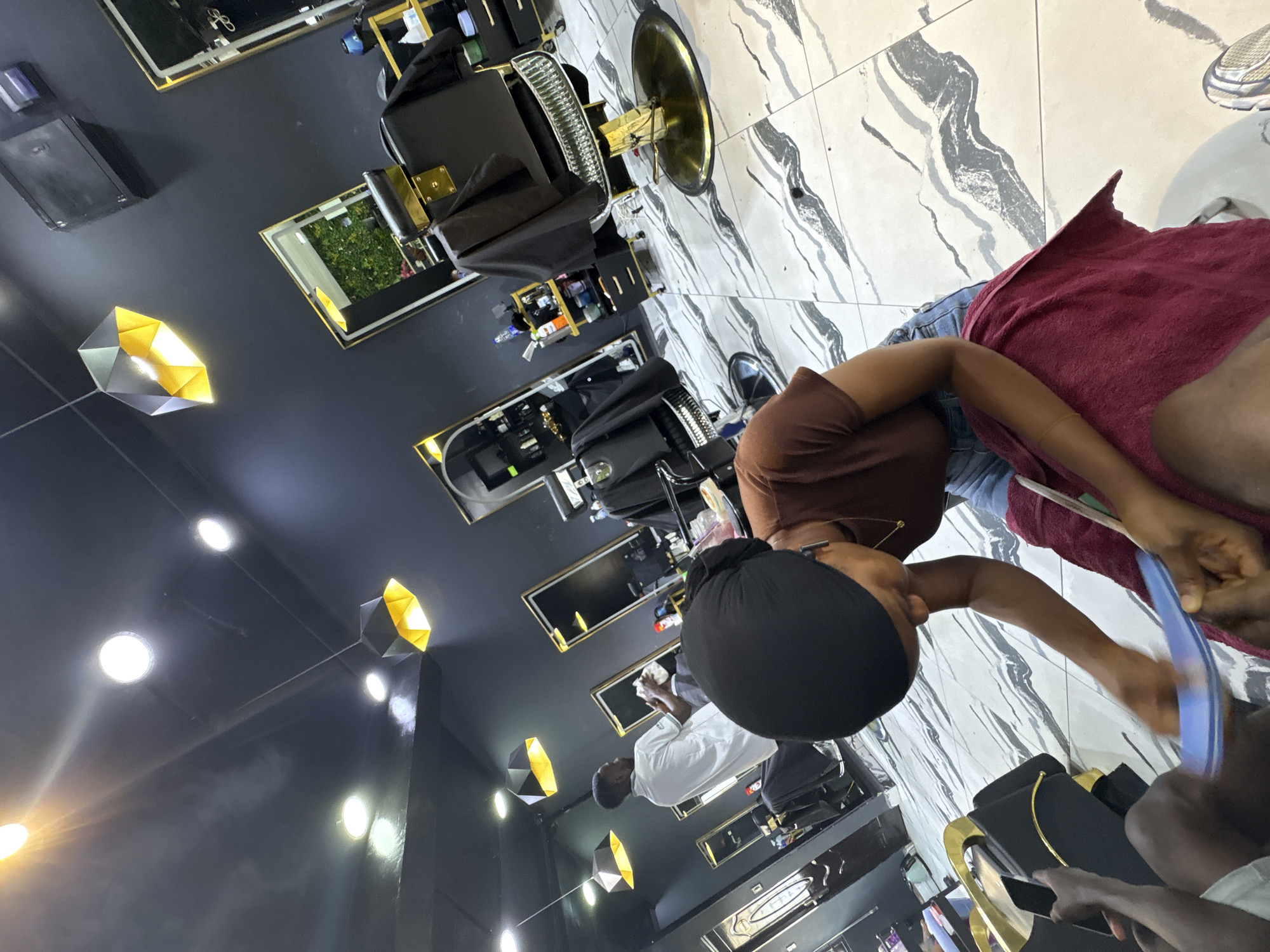 The image depicts a scene inside a business, possibly a salon, located in Lugbe, Nigeria. The floor is tiled with a white and gray marble pattern. A person with a black head covering is visible, wearing a brown shirt and blue jeans. They are bent over, seemingly working on someone's feet, which are covered by a reddish-brown towel. A blue object is in their hand, possibly a tool used for pedicures. In the background, a man wearing a white shirt stands near salon chairs. The salon has black walls and features geometric light fixtures, black salon chairs with gold trim, and rectangular mirrors.