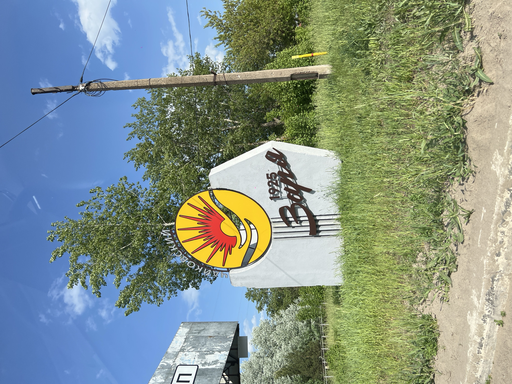 The image shows a sign marking the entrance to the city of Chaikovsky, Russia. The sign is a grey stone structure. It features a yellow sun with red rays and a smiling face, next to the inscription "1925 Чайковский". The sign is surrounded by green grass and trees. In the background, there's a utility pole against a blue sky with clouds. A fragment of another sign is visible in the lower left corner. The image appears to have been taken from a low angle.