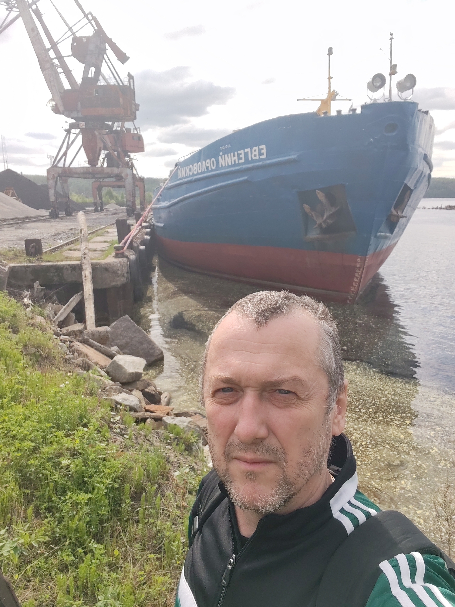 A selfie image captures a man standing on the rocky bank of a body of water in an industrial port setting, identified as Medvezhyegorsk, Russia. The man, with short, greying hair and a beard, is wearing a black and green track jacket and looks directly at the camera with a neutral expression.
Behind him, a large blue and red cargo ship is docked alongside a concrete pier. Its name, "Nnksoco Nnelbe," is visible in white capital letters on the hull, appearing mirrored and upside down due to the selfie camera's perspective (suggesting the actual ship name is "ЕВЛЕНН ОВОСККН"). Two prominent, rusty red industrial cranes, characteristic of a loading dock, are situated on the shore behind the ship, next to piles of dark material, likely coal or aggregate.
The scene is set under an overcast sky during daytime, providing soft, diffused light. Green grass and other vegetation grow along the rugged bank, which is strewn with rocks and debris. The water near the shore is clear, with a light, speckled appearance on its surface, possibly indicating shallow depth or a light-colored bottom. The distant background shows a treelined shore across the water.