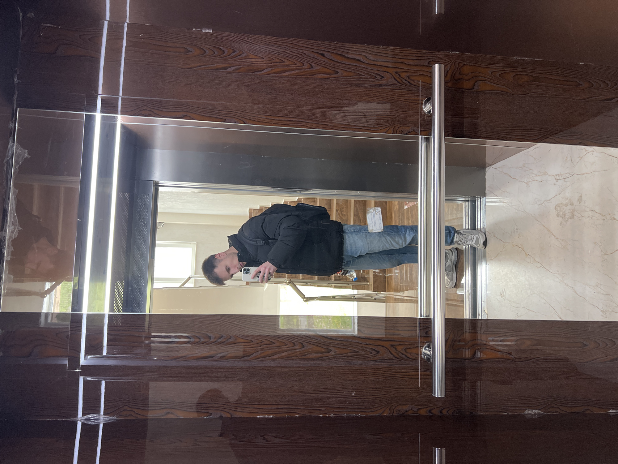 A man in a black jacket and jeans is reflected in a mirrored elevator door. He's holding a white cell phone. The elevator appears to be within a building in Burgas, Bulgaria, based on the provided information. The elevator doors have a wood-grain pattern on the outside and are stainless steel on the inside. The surrounding hallway appears to be finished with light-colored marble tile. There is a set of stairs visible in the reflection.
