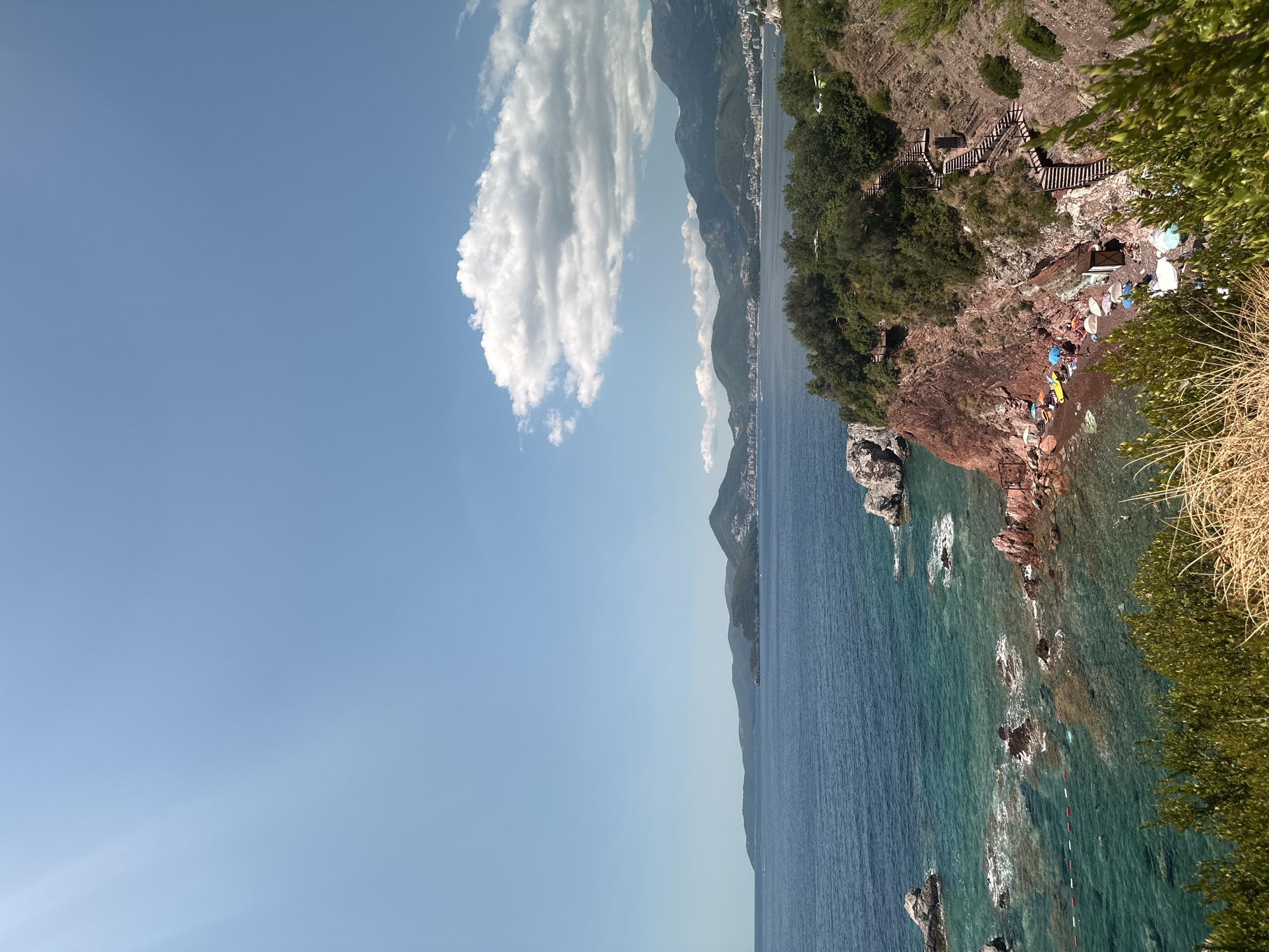 This is a high-angle, wide shot of a rocky coastline in Sveti Stefan, Montenegro. The sea, a vibrant mix of blues and greens, stretches to the horizon, where mountains dotted with buildings form a distant backdrop. The foreground is dominated by a lush green hillside, with dry grass in the immediate front. A small, secluded beach is nestled among the rocks, with several people visible sunbathing on towels and loungers under colorful umbrellas. Some kayaks are pulled up onto the beach. A wooden staircase winds its way down the steep, reddish-brown cliff face, providing access to the beach. The sky above is a clear blue, with a few fluffy white clouds adding visual interest. Sunlight streams in from the left side of the frame, casting subtle rays across the sky.