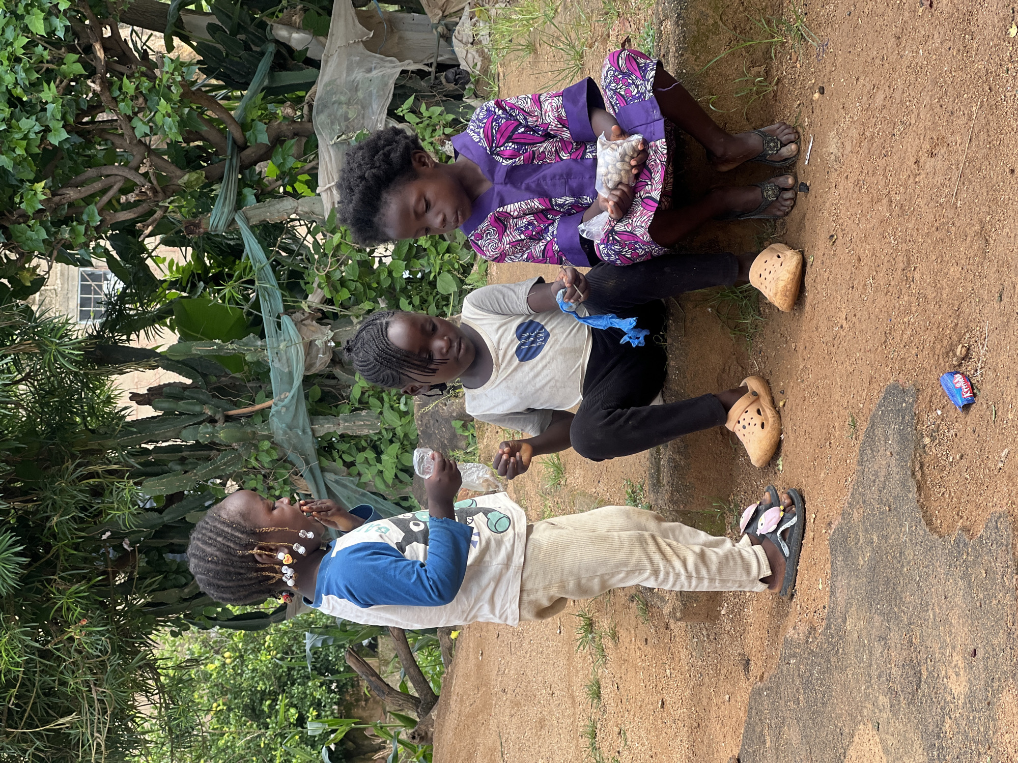 Three young children are gathered on a dirt path in a seemingly outdoor, urban environment. The child on the left, wearing a blue and white long-sleeved shirt and beige pants, stands with their back partially to the viewer and is holding a clear plastic bag with a small object inside. This child has braided hair adorned with colorful beads.
In the center, a child in a white t-shirt and black pants sits on a low concrete ledge. They are looking down at a blue plastic bag in their hands.
To the right, a child dressed in a vibrant purple and pink patterned outfit sits on the same ledge. This child is holding a clear plastic bag filled with what appear to be small nuts or seeds. Their hair is short and curly.
The ground is a reddish-brown dirt with sparse patches of dry grass. Behind the children, lush green foliage and trees create a natural backdrop, with some evidence of a building and a window visible through the leaves. The overall impression is that of a casual gathering of children in an outdoor space within a populated area, possibly in Jos, Nigeria, given the visual cues of vegetation and architectural style that are common in such locations. A discarded colorful wrapper is visible on the ground in the foreground.