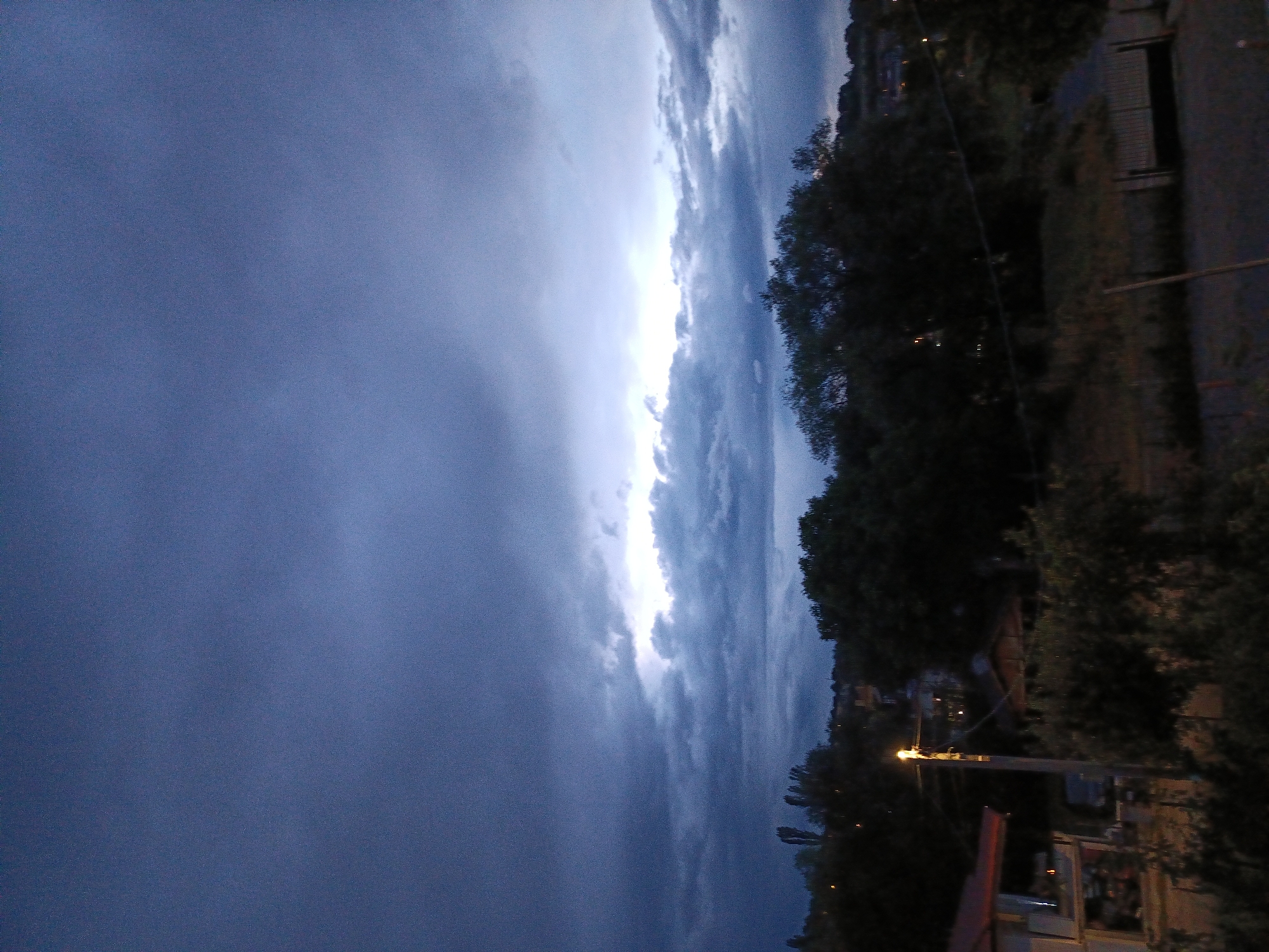 A twilight scene over Kralev Dol, Bulgaria, features a dark, cloudy sky with a bright band of light breaking through the clouds near the horizon. The foreground is dominated by silhouetted trees and vegetation. In the lower left, a building with a reddish roof and a lit lamppost on a pole are visible. The landscape appears to be a residential area with some houses dimly lit in the background, set against a backdrop of rolling hills covered in trees. The overall atmosphere is moody and subdued, indicative of approaching or receding daylight.