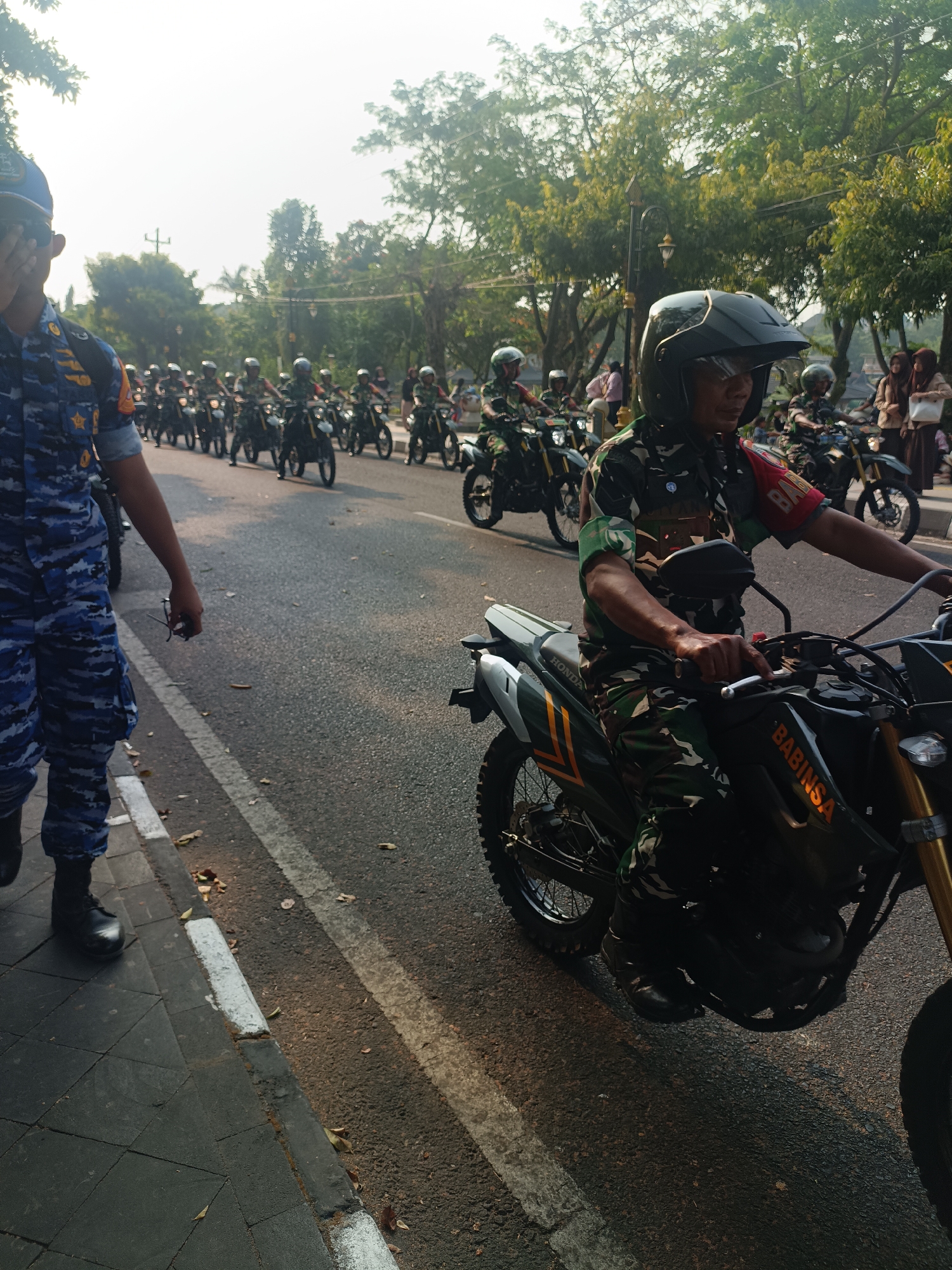 In the city of Sleman, Indonesia, a group of Indonesian military personnel in camouflage uniforms are participating in a motorcade. The image captures a procession of motorcycles, with several riders in focus. In the foreground, a soldier wearing a black helmet and camouflage uniform is riding a dark-colored motorcycle with "BABINSA" emblazoned on its side. Another individual in a blue camouflage uniform and a blue cap is seen walking on the sidewalk to the left, partially covering their face with their hand. Behind the motorcycles, trees line the street, and a bright, hazy sky suggests it is daytime. The road is paved, with white lines marking the lanes. In the background, other riders on motorcycles are visible, along with a few civilians observing the event.