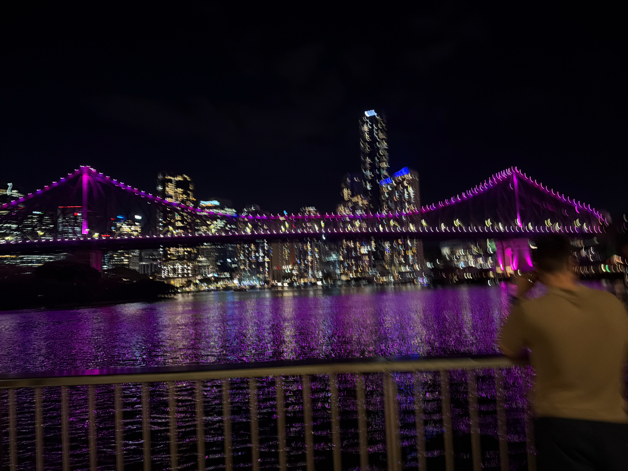 An out-of-focus nighttime scene depicts the Story Bridge in New Farm, Australia, illuminated with vibrant purple lights. The bridge spans a body of water that reflects the city lights. In the foreground, a person stands behind a railing, facing away from the camera, appearing to take a photo or video with their phone. The background showcases a blurred cityscape with numerous buildings and lights. The overall impression is one of a lively urban environment at night.