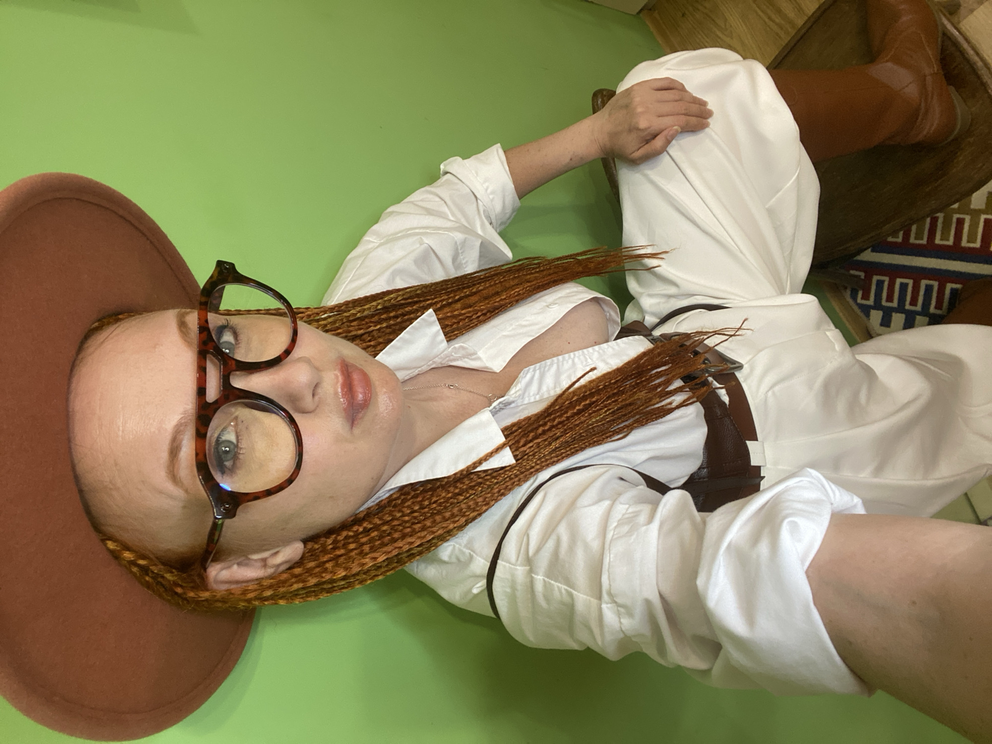 This is a high-angle, full-body, portrait-orientation photograph of a young woman with long, braided, reddish-brown hair. She is wearing a white button-down shirt, a brown belt, and brown leather boots. Her hair is styled in thin braids that fall over her chest. She is wearing tortoise-shell glasses and has on light makeup, including lip gloss. The woman is reclining on a surface that appears to be a chair or couch, with her body positioned diagonally across the frame. Her left arm is bent at the elbow, with her hand resting on her torso, while her right arm is extended upwards and out of the frame.
The background is a solid, bright green wall, with a wooden floor visible in the upper right corner. A dark wooden chair and a patterned rug are also present in the background. The lighting appears to be artificial, suggesting an indoor setting. The overall mood of the image is relaxed and casual, possibly a candid shot or a posed photograph in a home environment. There is no visible text in the image. The location context is Sochi, Russia.