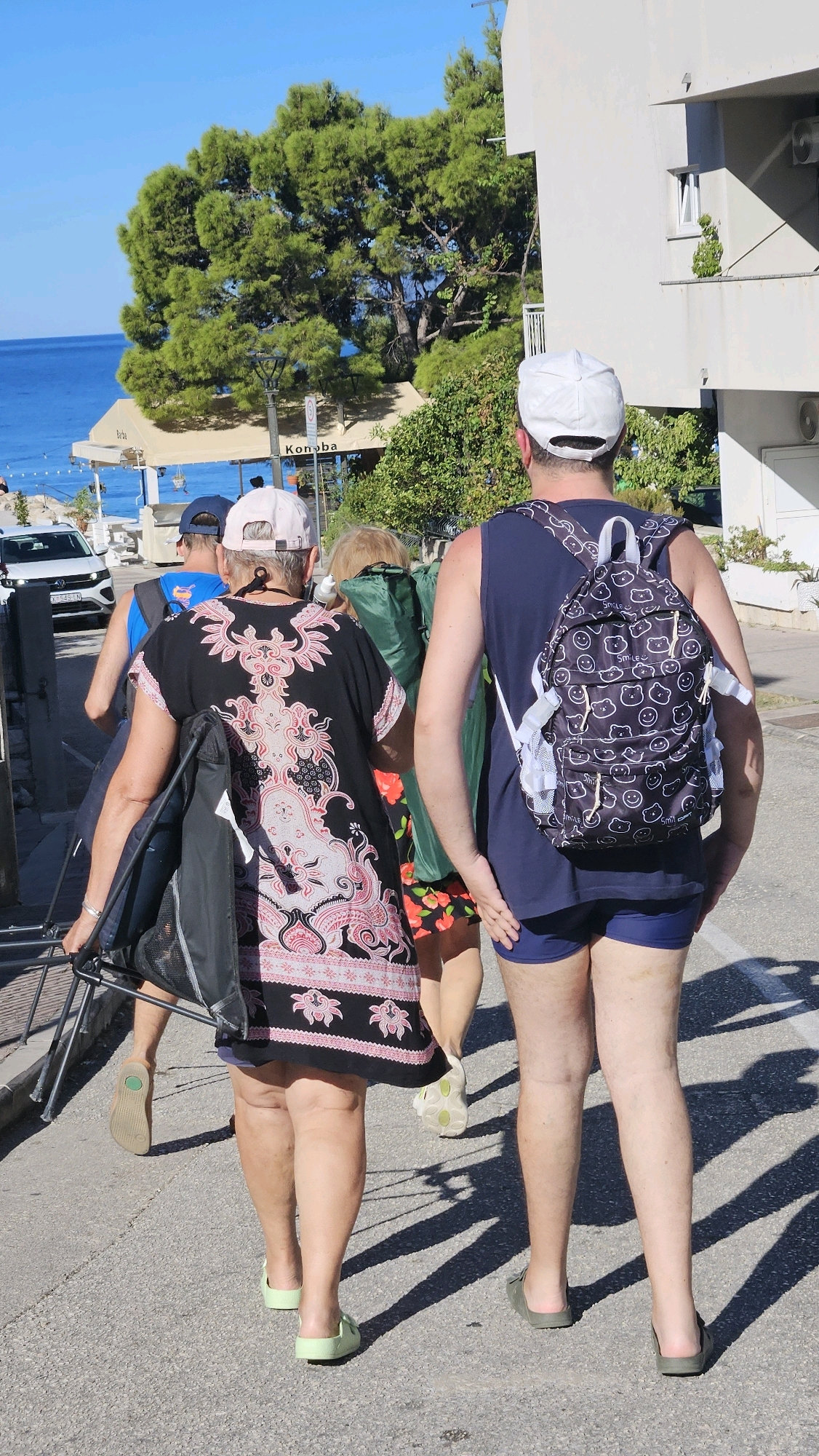 In Općina Tučepi, Croatia, on a sunny day, four individuals walk along a paved street. The foreground features two people walking away from the camera. The person on the left is wearing a black dress with a pink and white patterned design, light green sandals, and is carrying a disassembled black chair. The person to their right is a man wearing a navy blue tank top, navy blue shorts, and dark green slides, carrying a backpack with a white and black smiley-face pattern. Behind them, a person in a blue shirt and a blue cap walks with a backpack, and another person with blonde hair wears a green shirt and a floral skirt. A white car is visible in the background, parked on the left side of the street, with the blue sea and lush green trees beyond it. A white building with windows and air conditioning units stands to the right. The ground is asphalt, with distinct shadows cast by the sun.