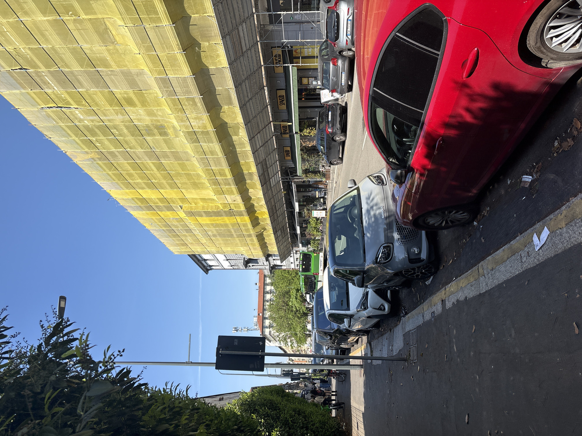 A street scene in Milan, Italy. Along the right side of the street, parked cars line the curb, including a red car at the forefront, followed by a gray Smart car and other vehicles extending into the distance. A line of shops fronts the street on the opposite side, partially obscured by scaffolding draped in yellow protective material. A street sign and light pole stand on the left. Green foliage is visible in the lower left corner. People are present further down the street. The sky above is blue.