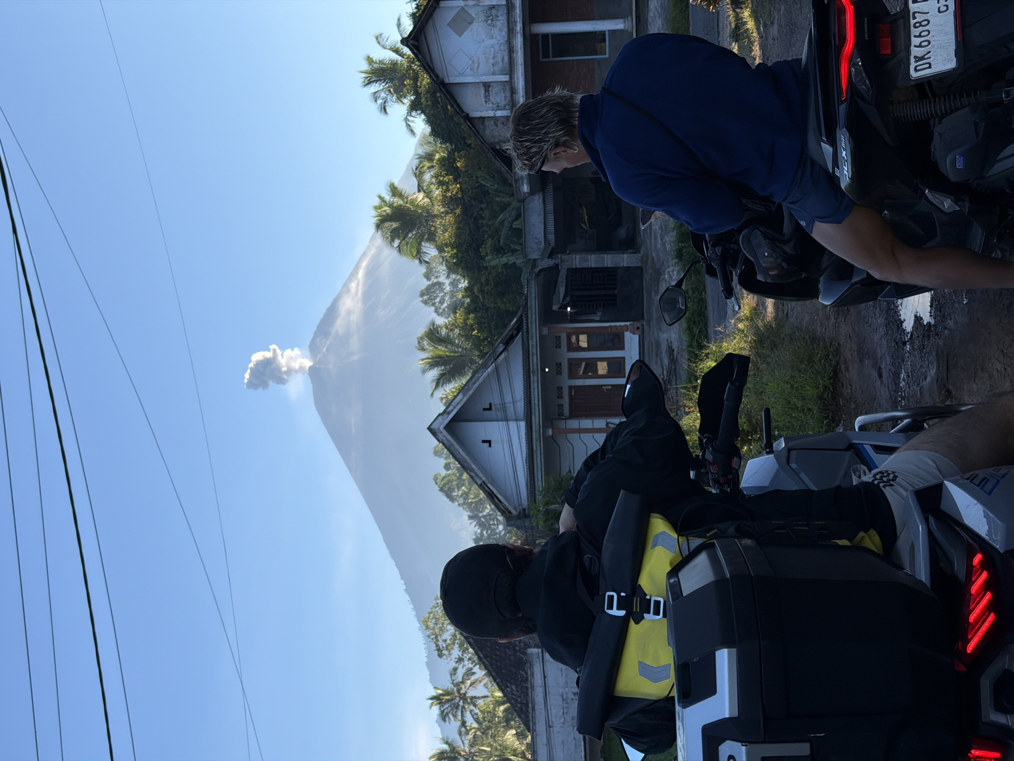 In Sidomulyo, Indonesia, two individuals are seated on motorcycles, facing away from the camera, observing a volcano erupting in the background. The volcano is spewing a plume of ash into the clear blue sky. In the foreground, on the left, a person wearing a black cap and jacket is on a motorcycle with a yellow bag attached to the back. On the right, another person in a blue jacket and dark shorts is on a separate motorcycle, with a visible license plate that reads "DK 6687 F". Between the motorcycles, traditional Indonesian houses with tile roofs are visible, surrounded by lush green vegetation and palm trees. The ground appears to be dirt and wet in places. The scene is illuminated by natural daylight, suggesting it is either early morning or late afternoon.