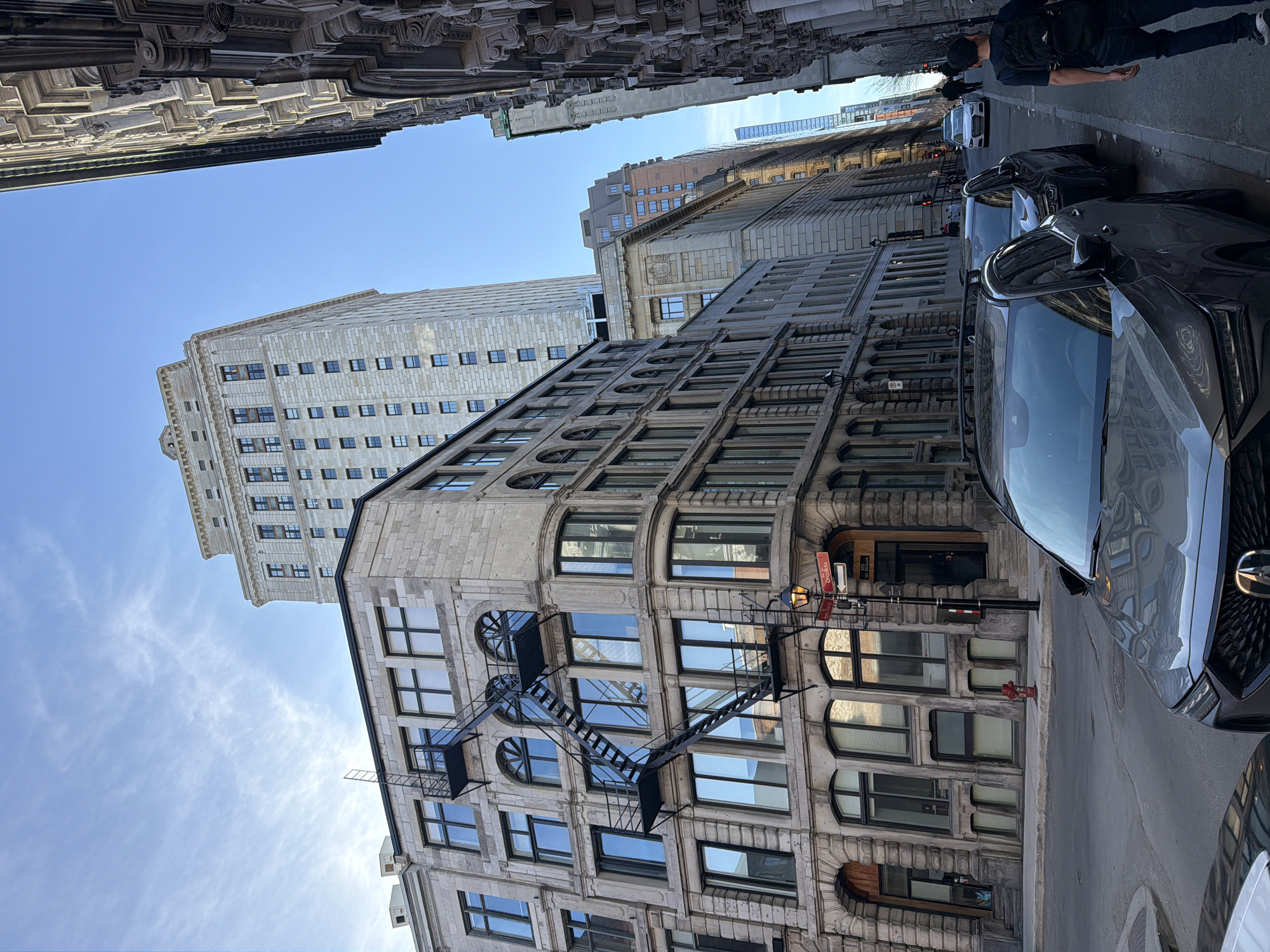 The image captures a street scene in Montreal, Canada. The camera is pointed upwards, framing the architecture of buildings on either side of the street. A taller building of light stone is visible in the background with rectangular windows. An adjacent building shows arched windows and a fire escape. Modern cars line the right side of the street. A person walks along the sidewalk to the right of the frame. Overhead, architectural details of surrounding buildings are visible. A clear, blue sky can be seen between the buildings.