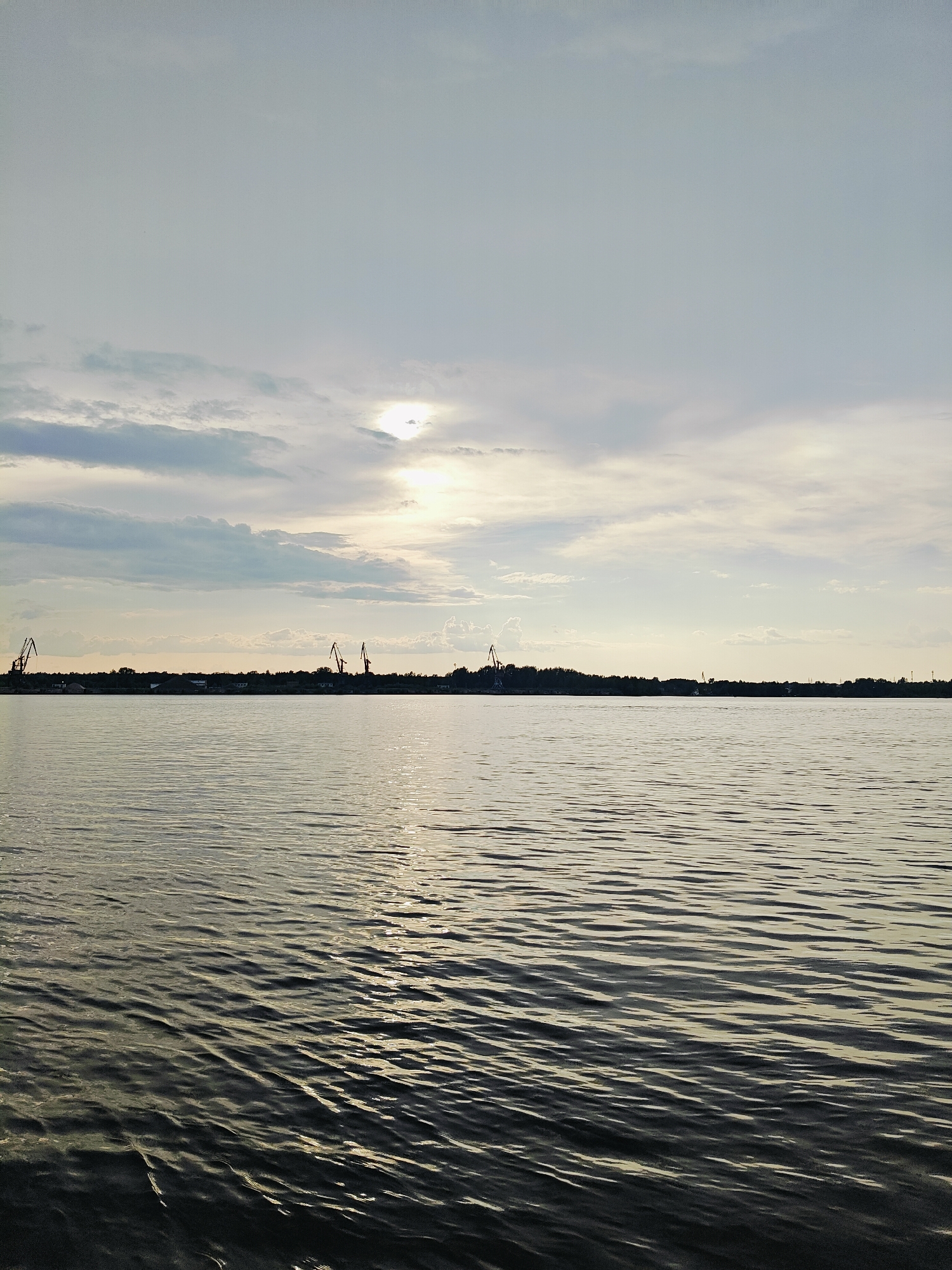 The image depicts a wide body of water, likely a river or reservoir, with gentle ripples reflecting the overcast sky. On the horizon, a tree line separates the water from the sky, with several industrial cranes visible against the lighter sky. The scene suggests an industrial or port area, possibly along the Kama River in Chaikovsky, Russia, given the presence of such infrastructure and the characteristic landscape of the region. The lighting indicates it is likely late afternoon or early morning, with diffused sunlight breaking through the clouds. There are no people or animals visible in the frame.