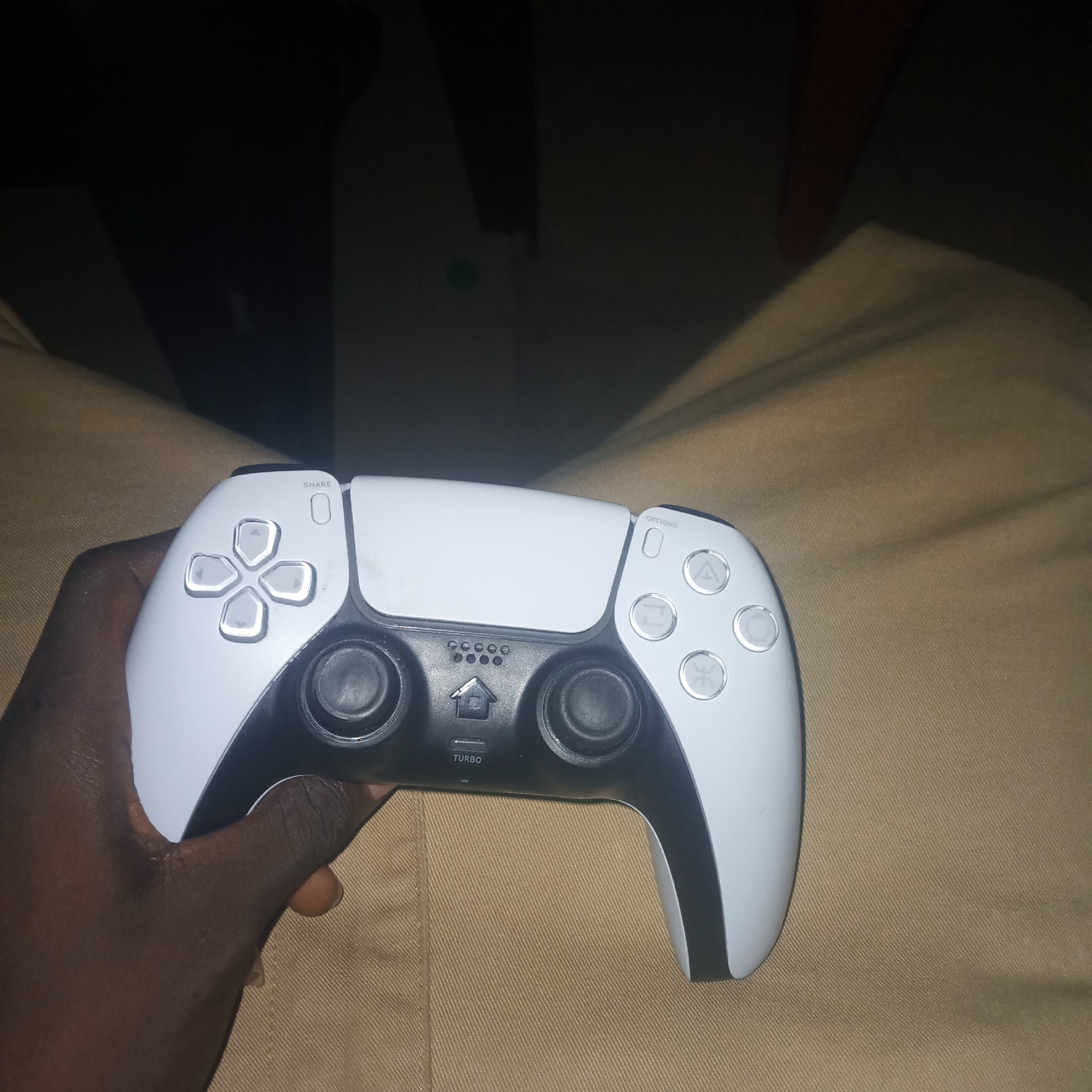A left hand is holding a white PlayStation 5 DualSense controller. The controller is oriented horizontally, with the directional buttons on the left and the face buttons on the right. The directional buttons are illuminated with white light. The hand is positioned on the left side of the controller, with the thumb resting on the left analog stick. The background is a dark, out-of-focus environment. The image is taken from a slightly elevated angle, looking down at the controller and hand.