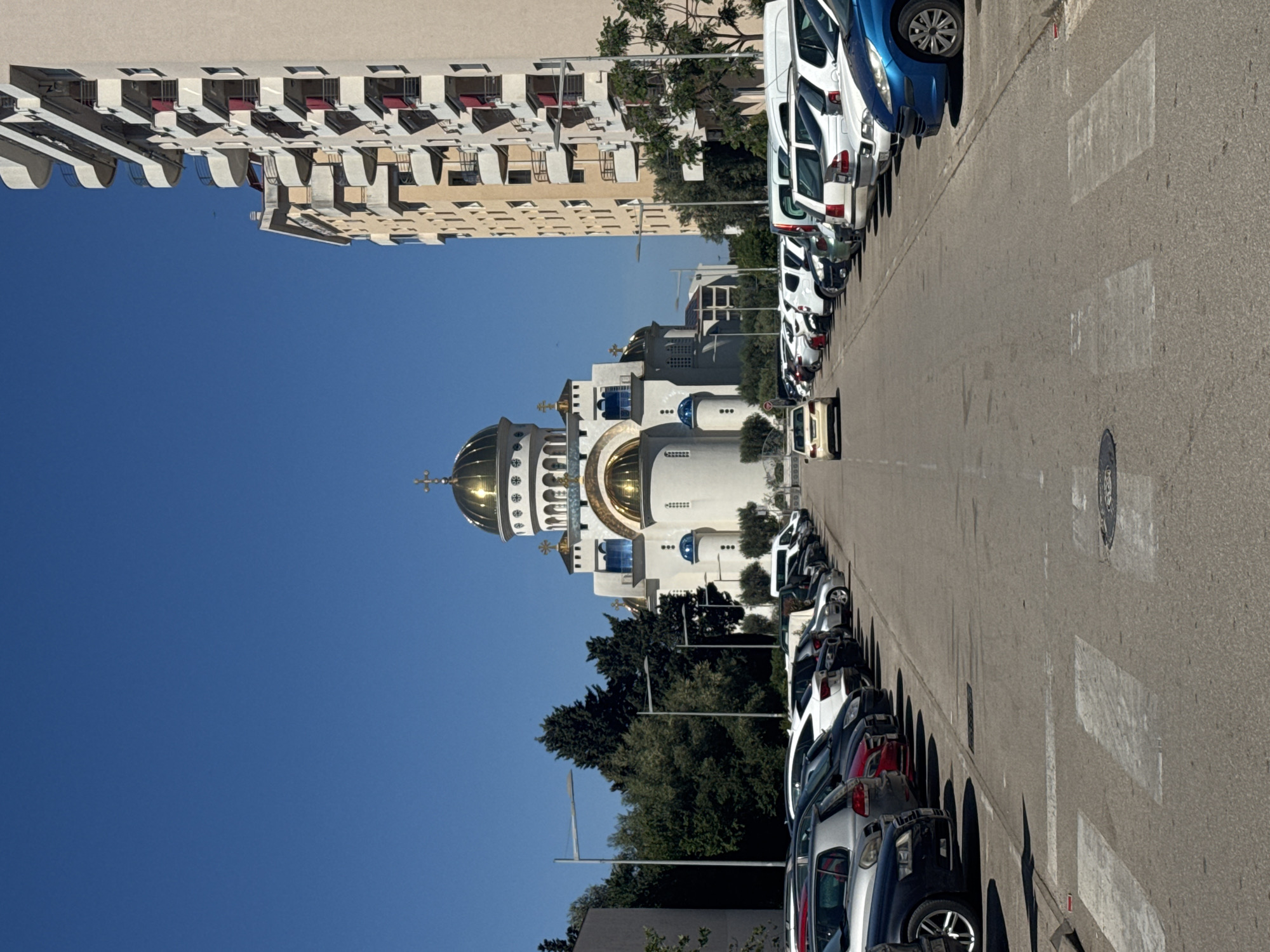The image shows a street scene in Bar, Montenegro. A line of parked cars lines the right side of the asphalt road, their shadows cast on the pavement. On the left side, a large, white Orthodox church with golden domes stands prominently against a clear blue sky. Behind the church, there are green trees. Further in the background, apartment buildings can be seen. The image is taken during the day, with bright sunlight illuminating the scene.