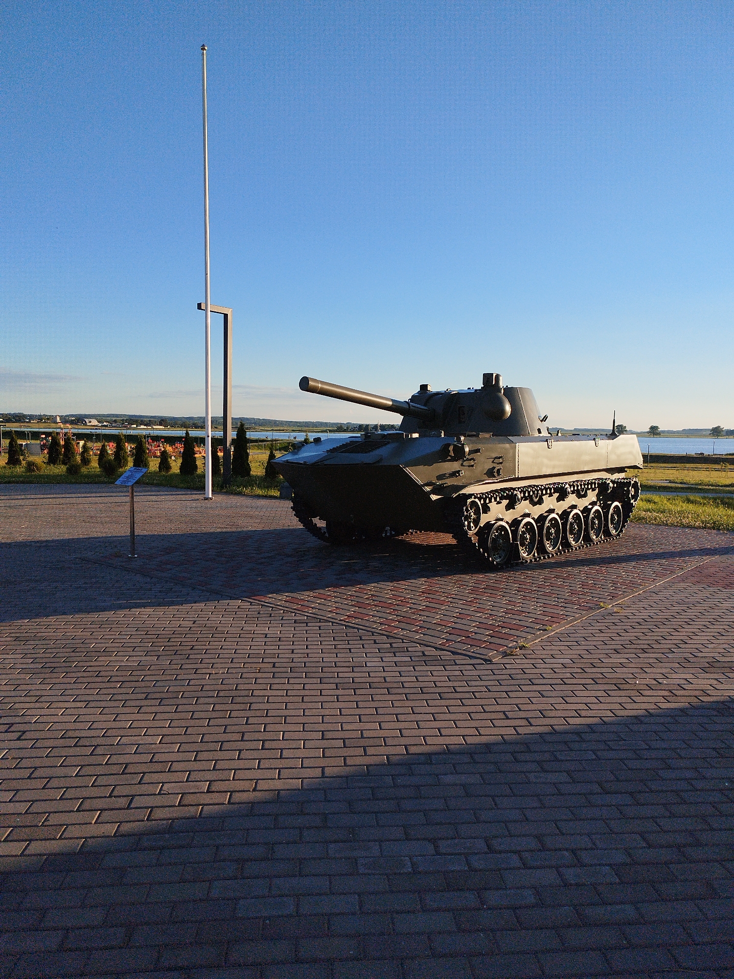 An exterior, eye-level, wide shot captures a dark green military tank on a paved area under a clear blue sky. The tank is positioned in the lower right quadrant of the frame, with its tracks and turret clearly visible. In the background, a body of water is visible, with a distant shoreline featuring trees and some buildings. To the left of the tank, a tall flagpole stands prominently, with a small informational plaque at its base. The paved area in the foreground is made of interlocking bricks, with shadows cast from the objects in the scene. The overall lighting suggests it is either early morning or late afternoon, with the sun casting long shadows.