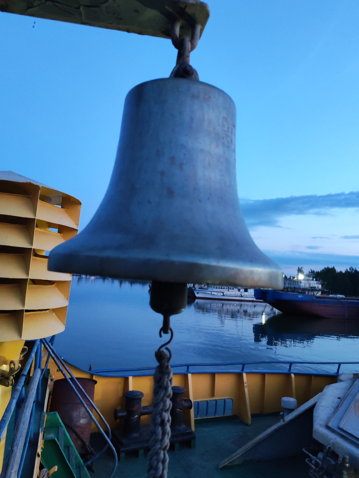 The image depicts a close-up view of a ship's bell hanging from a metal fixture against a twilight sky. The bell is large, metallic, and shows signs of weathering. To the left, a portion of a yellow structure with a honeycomb-like pattern is visible. Below the bell, a rope is attached. The background shows a body of water reflecting the sky and distant vessels. The deck of the ship, with yellow railings and metal posts, is also in the foreground. The scene suggests a nautical setting, possibly at dusk or dawn. The location is specified as Bol'shie Mkhi, Russia.