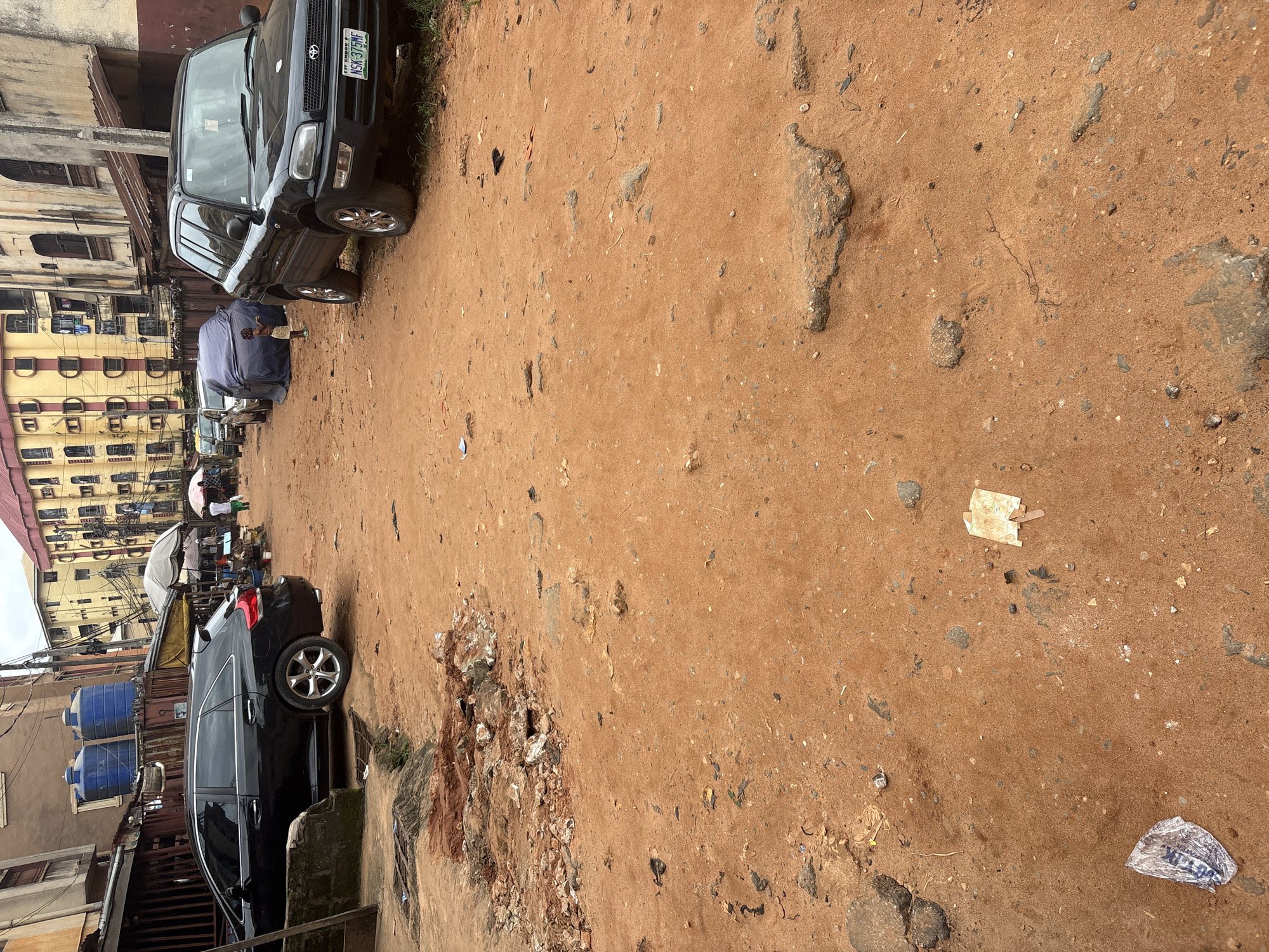 In Onitsha, Nigeria, a dirt road is filled with debris, including broken concrete and scattered trash. Two black SUVs are parked on the side of the road. One SUV, a Toyota RAV4 with license plate NSK-375MF, is closer to the camera. The other SUV is further down the road. Several buildings line the street, their architecture suggesting a dense urban environment. A child stands near a car covered with a blue tarp. In the background, market stalls are set up, with a few people visible among them. The ground is uneven and appears to be under construction or in disrepair, with piles of red soil and rocks creating an obstacle course.