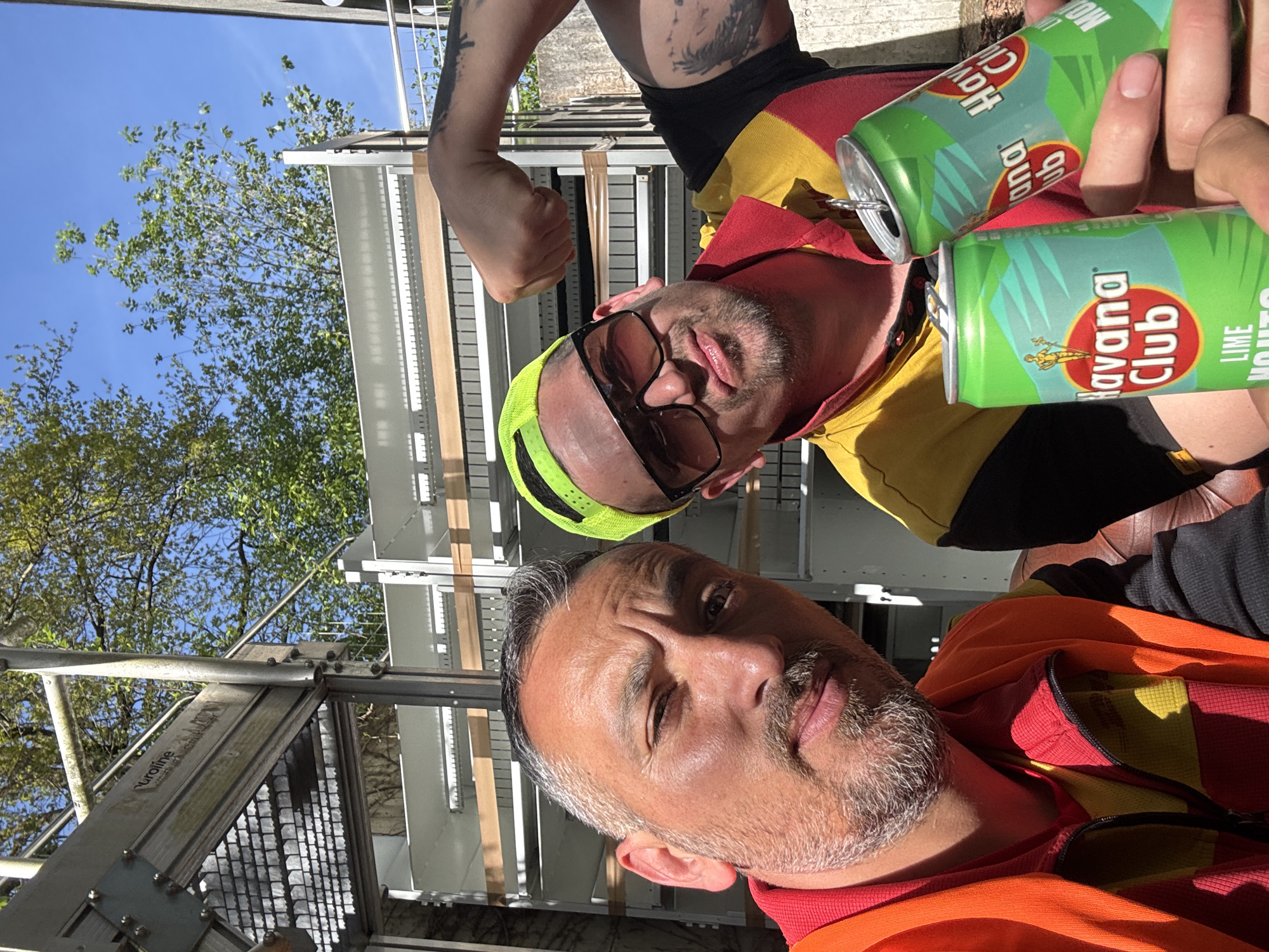 This outdoor, bright daylight image taken in Bietigheim-Bissingen, Germany, features two men posing for a casual photo. In the foreground, a man with a graying beard and mustache wears an orange and red striped long-sleeved shirt/jacket. He looks towards the camera with a slight smirk, his right eye partially squinting, possibly due to the bright sun. Behind him, a second man wears a yellow, red, and black striped polo shirt, dark sunglasses, and a neon yellow visor. He flexes his tattooed left arm and holds two green cans labeled "Havana Club LIME MOJITO." One of the cans is open with a straw inserted.
The setting includes a clear blue sky with bright sunlight, green-leafed trees, and industrial-style metallic structures, possibly scaffolding or part of a modern building. Faint text reading "uruline" is visible on one of the metal beams in the background. The scene conveys a relaxed, social atmosphere under sunny weather conditions, likely during midday or early afternoon.