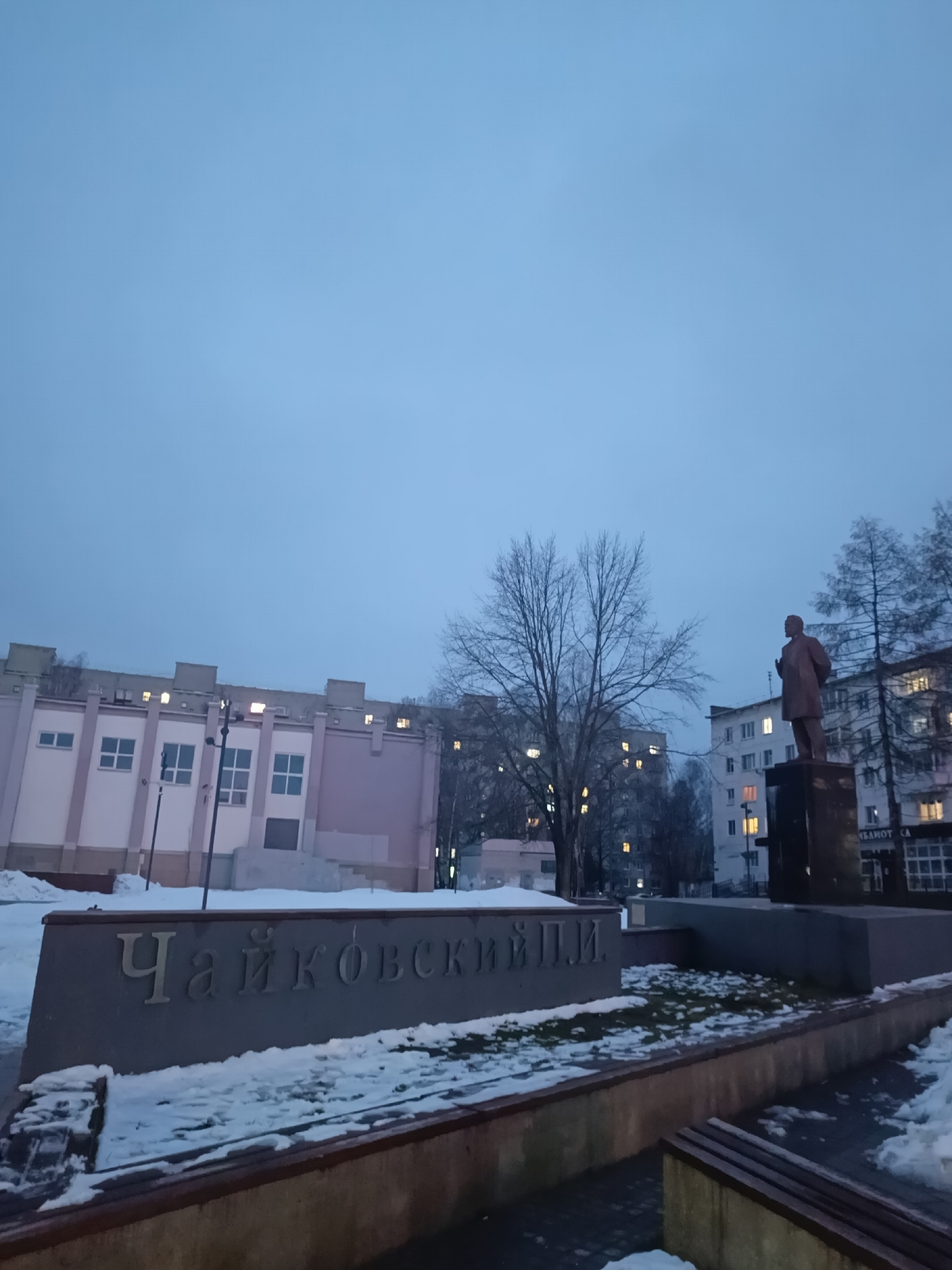 The image depicts a public square in the city of Chaikovsky, Russia, likely taken in the early morning or late evening due to the muted light. A statue, potentially a historical figure, stands on a pedestal to the right of the frame. In the foreground, a low wall or platform bears the name "Chaikovsky" in Cyrillic letters. Patches of snow are visible on the ground and on the wall, indicating a cold season. Buildings, some with lit windows, form the backdrop to the square. Bare trees are interspersed between the buildings and the statue, contributing to the wintry atmosphere.