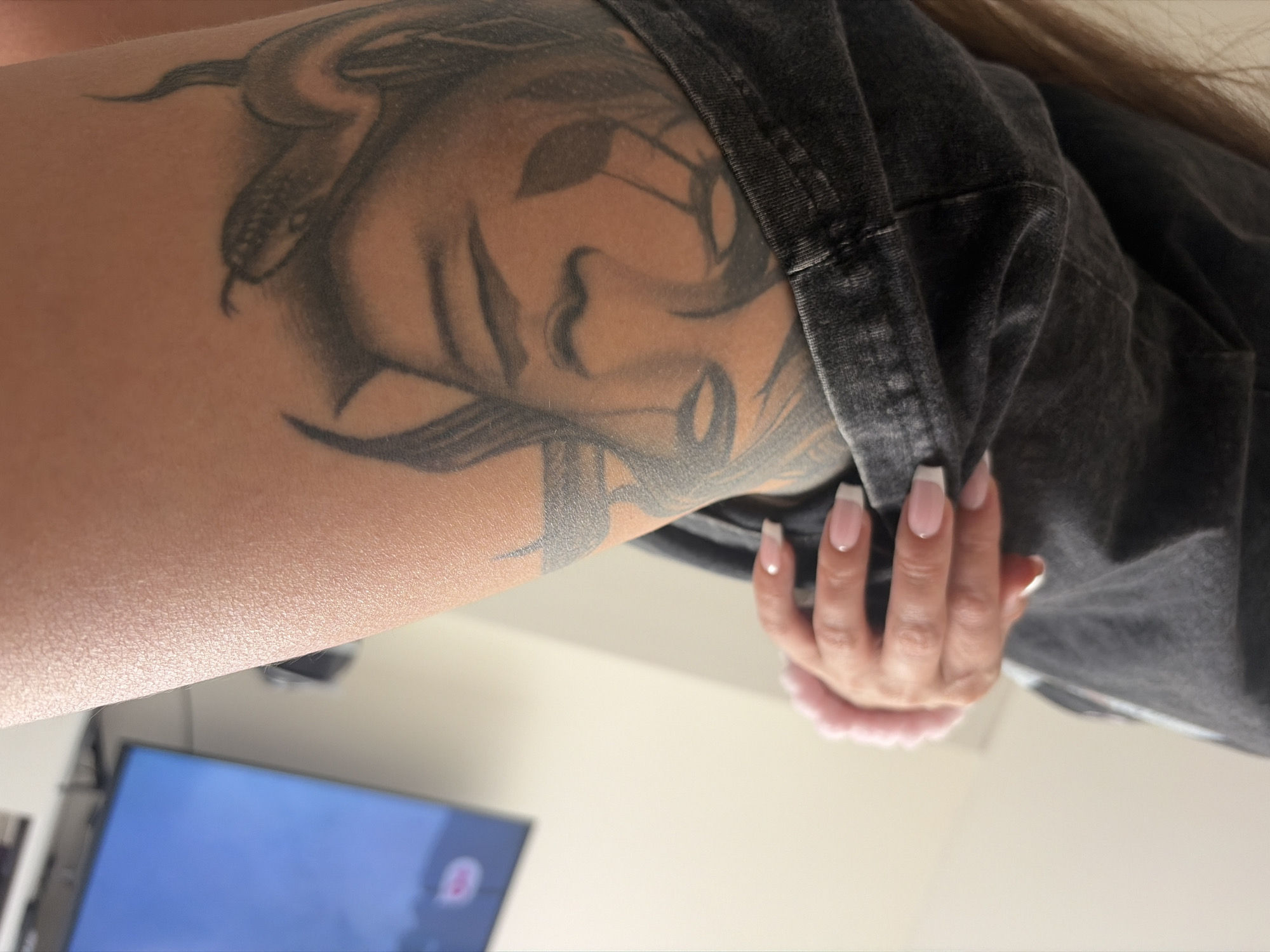 The image depicts a close-up view of a person's upper arm and hand. The arm features a tattoo with a snake motif interwoven with a woman's face. The hand, adorned with manicured nails with a French tip design, is holding a grey garment. In the background, a television screen and a light-colored wall are visible. The location in Saint Petersburg, Russia, is not discernible from the provided image.
