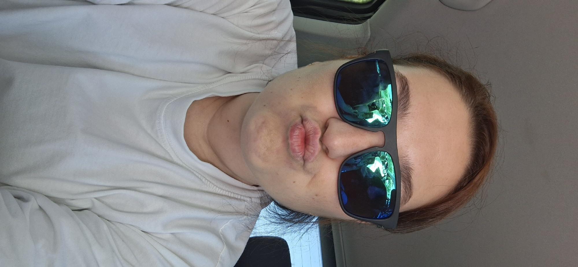 A person with fair skin and reddish-brown hair is posing for a selfie while wearing sunglasses. The sunglasses reflect a blue and green hue, obscuring the person's eyes. The person has pursed lips. They are wearing a white long-sleeved shirt. The photo appears to have been taken inside a vehicle, judging by the interior paneling and the light visible in the sunglasses' reflection. It is not possible to determine the exact location in Chaikovsky, Russia, from this image alone.