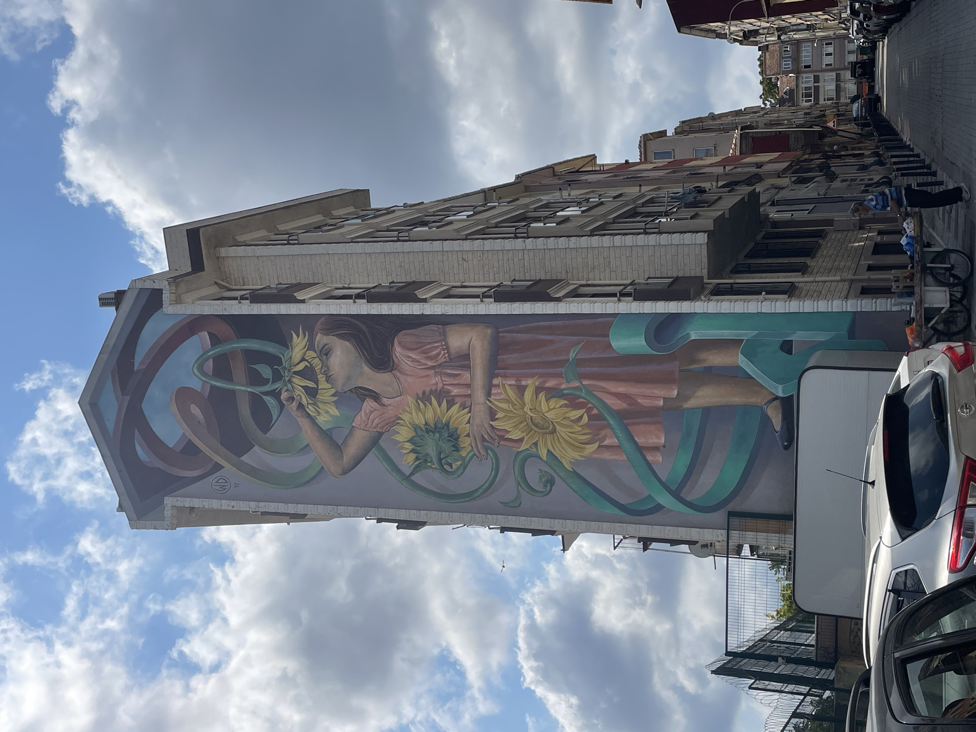The image captures a street scene in Kadıköy, Turkey, dominated by a large mural on the side of a tall building. The mural depicts a young girl in a pink dress holding sunflowers, with teal-colored ribbons weaving around her. The building itself is modern with multiple floors and balconies. In the foreground, several cars are parked on the cobblestone street. To the right of the mural, a man wearing a striped shirt and dark pants stands beside a cart with various items on it. The street is lined with more buildings and parked cars, suggesting a residential or mixed-use urban area. The sky above is partly cloudy.