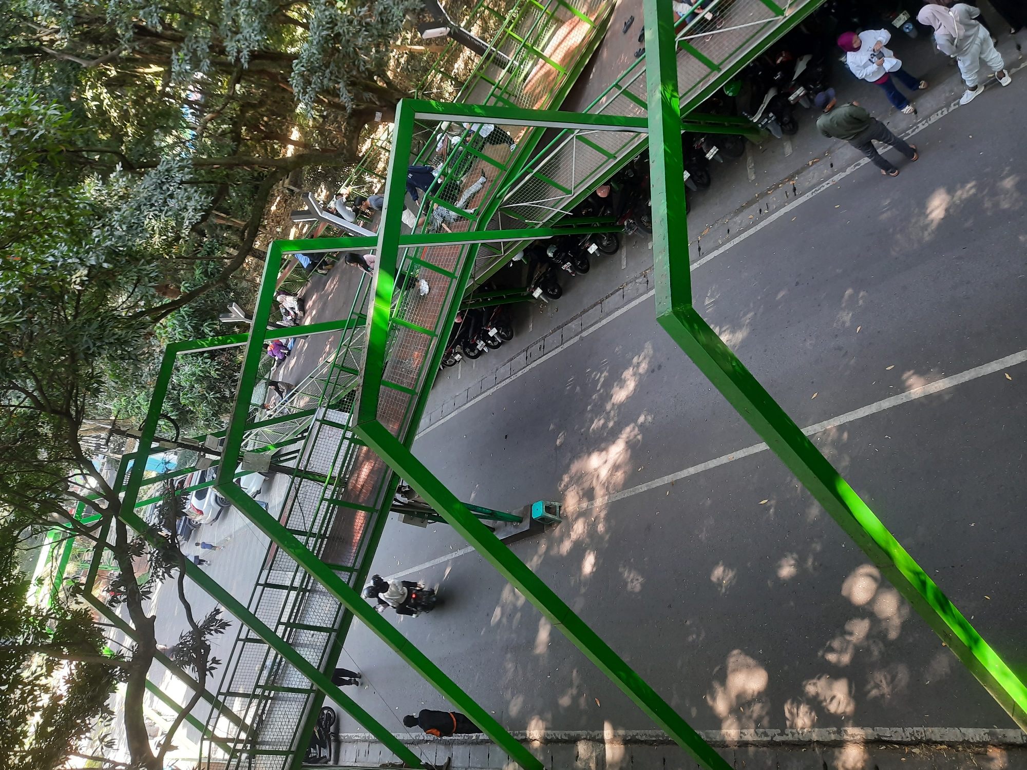The image depicts a pedestrian bridge with a green metal frame over a street in Bandung City, Indonesia. The bridge is elevated and has a walkway with metal railings. Below the bridge, several motorcycles are parked along the side of the road, which has a white dividing line. People are visible on the bridge and the sidewalk, with some walking and others standing. Trees with lush green foliage overhang the scene, casting dappled shadows on the ground. The overall impression is of an urban environment with pedestrian infrastructure integrated with the road and surrounded by nature.