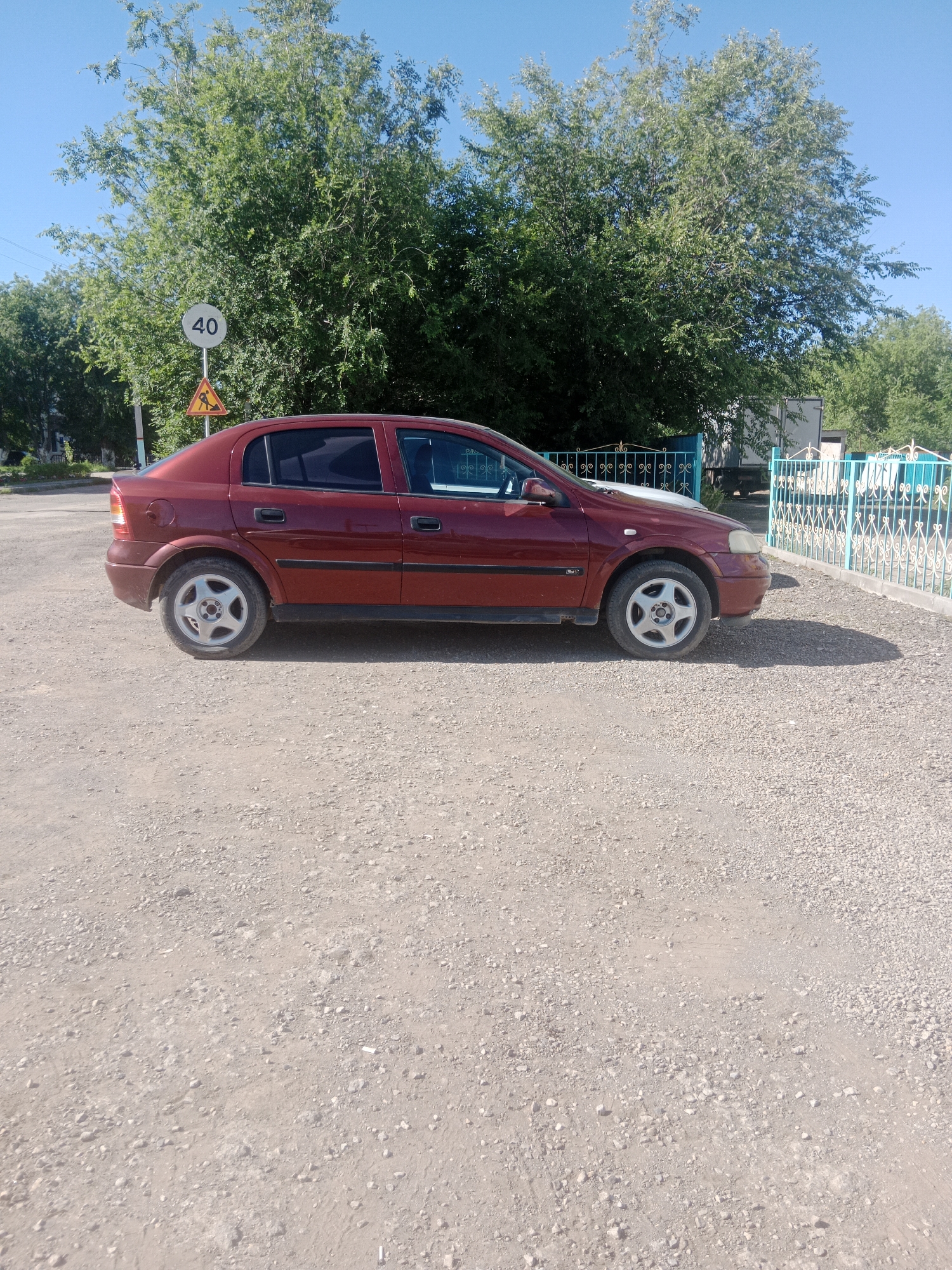 In Qobda, Kazakhstan, a red sedan is parked on a gravel lot. The car is positioned on the left side of the image, facing right, with its passenger side visible. The vehicle appears to be a model from the early 2000s, possibly an Opel Astra. Behind the car, a vibrant green tree line dominates the background, with lush foliage forming a dense canopy against a clear blue sky. To the right of the trees, a turquoise decorative metal fence with ornate scrollwork separates the gravel lot from what appears to be a property or pathway. A traffic sign indicating a speed limit of 40 is visible on a pole near the car, accompanied by a yellow triangular warning sign featuring a pedestrian symbol. The ground is covered with a mixture of small stones and dirt, suggesting an unpaved area. The scene is lit by bright sunlight, casting shadows from the car and the fence onto the gravel.