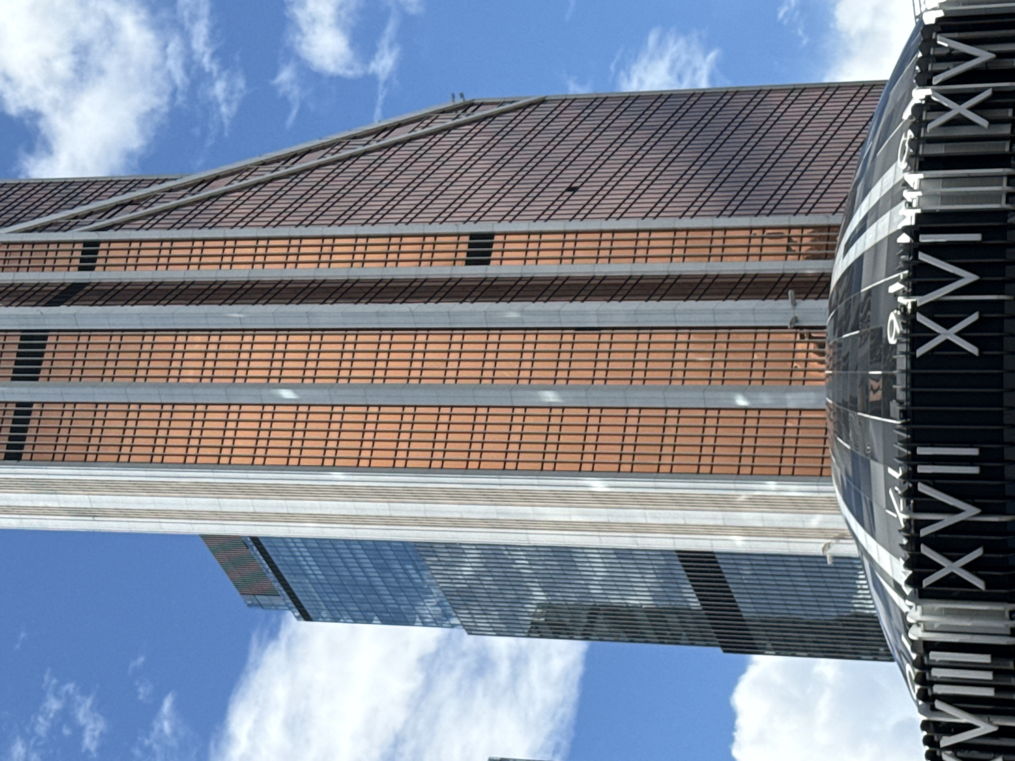 The image shows a close-up, low-angle view of the Moscow International Business Center, also known as Moscow City, in Moscow, Russia. The dominant features are two skyscrapers. The skyscraper on the right is the Neva Towers, characterized by its copper-colored glass facade with a grid pattern and vertical silver accents. It has a distinctive angled shape on its right side. To the left of the Neva Towers is another skyscraper with a predominantly blue-tinted glass facade, reflecting the sky and clouds. The foreground features the distinctive domed roof of a building within the complex, adorned with Roman numerals that appear to read "XVIII" and "XIV" at the bottom. The sky is blue with scattered white clouds. The image captures the modern architecture of Moscow City on a clear day.