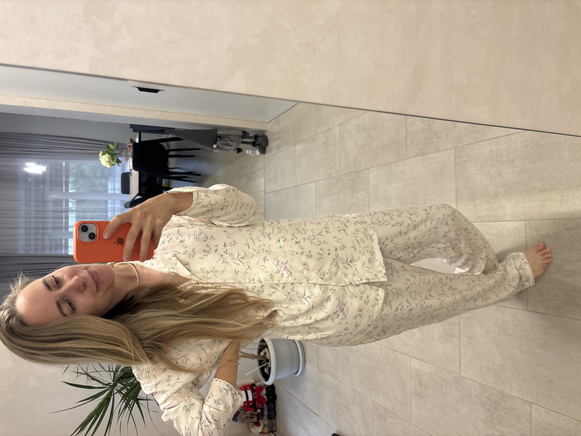 A young woman with blonde hair takes a selfie in a room. She is wearing a white pajama set with a delicate floral pattern. The pajama top has a collar and buttons down the front, and the sleeves have a ruffled cuff. The pajama bottoms are loose-fitting. She is holding an orange smartphone in her right hand, which is reflecting in the mirror. Her left arm is resting on her left leg.
The setting appears to be a home, possibly a bedroom or living area, with light-colored tiled flooring. In the background, a mirror reflects the room, showing a dining area with chairs and a table, and a window with grey curtains. There is a potted plant with long green leaves to her left. The overall mood is casual and relaxed. The lighting suggests it could be daytime. There is no visible text in the image.