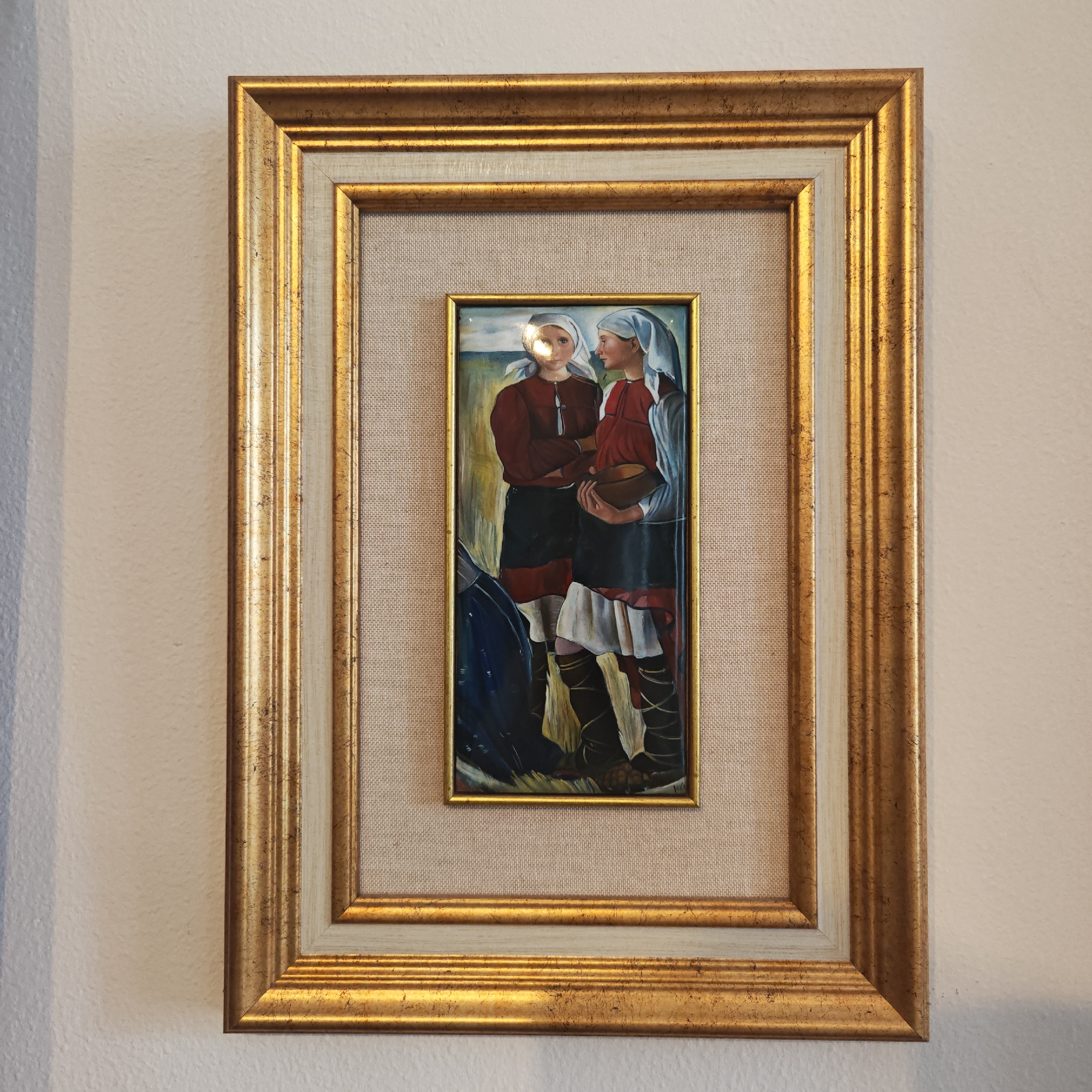 A framed painting hangs on a light-colored wall. The frame has a gold and off-white double border. The painting itself is a vertical rectangle depicting two women in traditional clothing. Both women wear white headscarves and red blouses. One woman is holding a wooden bowl. The setting appears to be a field or rural landscape. There are no visible indicators to confirm the location in Rostov, Russia.