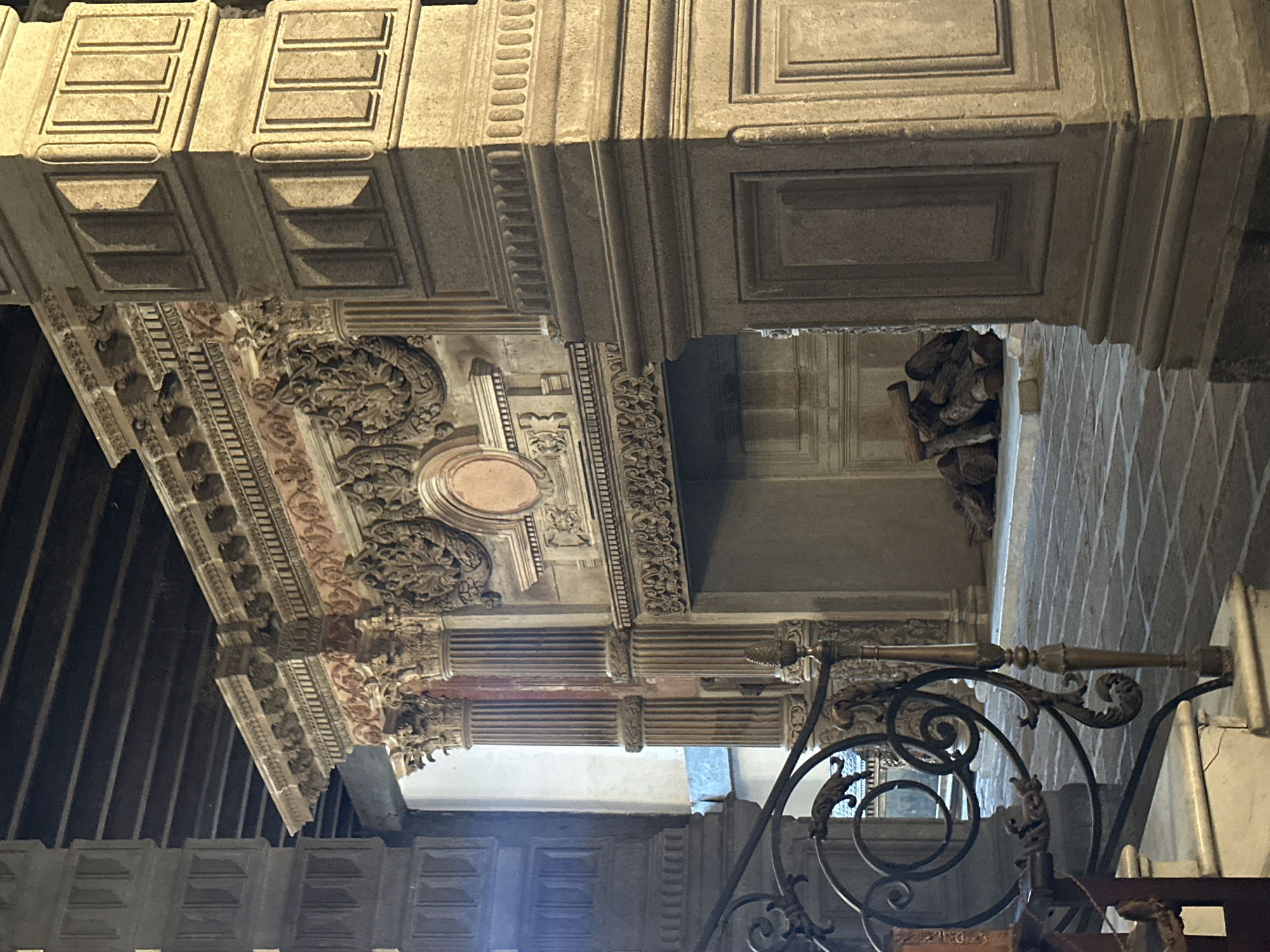 The image depicts an ornate, elevated architectural structure with heavily decorated elements. The structure features detailed carvings, including floral motifs, moldings, and panels with geometric patterns. There are columns and what appears to be coffered ceilings with recessed panels. The color scheme is primarily beige and brown. The style suggests a historical or religious building interior. A metal railing with decorative scrollwork is in the foreground. The location is most likely the Parish of the Body of Christ in the city of Tlalnepantla, Mexico. There are no people visible in the image. The environment is indoors, likely a church interior.