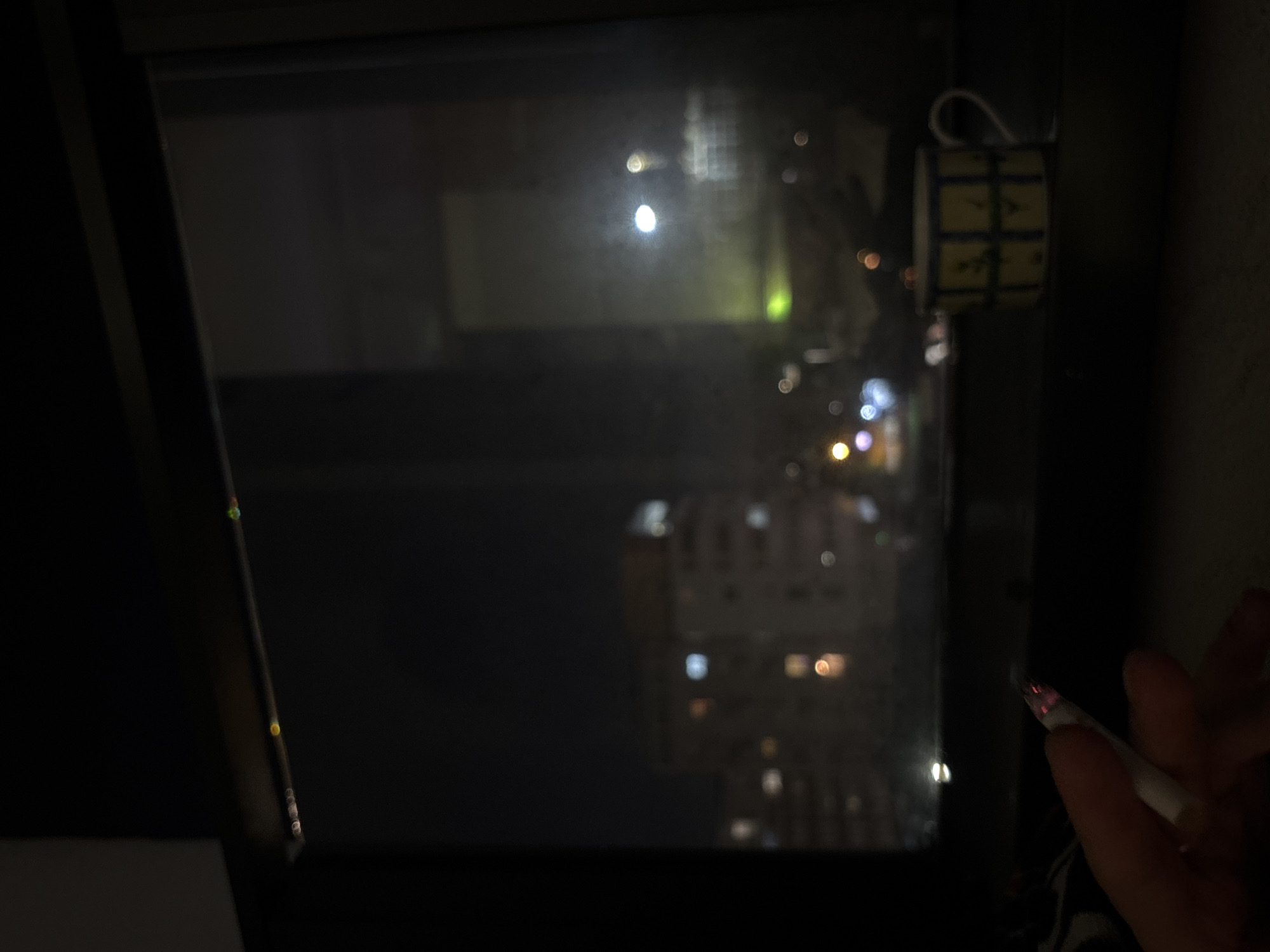 A dark, low-light photograph shows a person holding a lit cigarette in front of a window, with an out-of-focus cityscape visible through the glass. The window is in a dark room. The cityscape suggests a night scene in a dense urban area. The building lights appear blurred and distant. A small, cylindrical object with a grid pattern hangs near the top right corner of the window frame. The person's hand and the lit cigarette are closest to the camera, and are in the lower right corner of the image. There is not enough information to positively identify the location as Belgrade, Serbia.