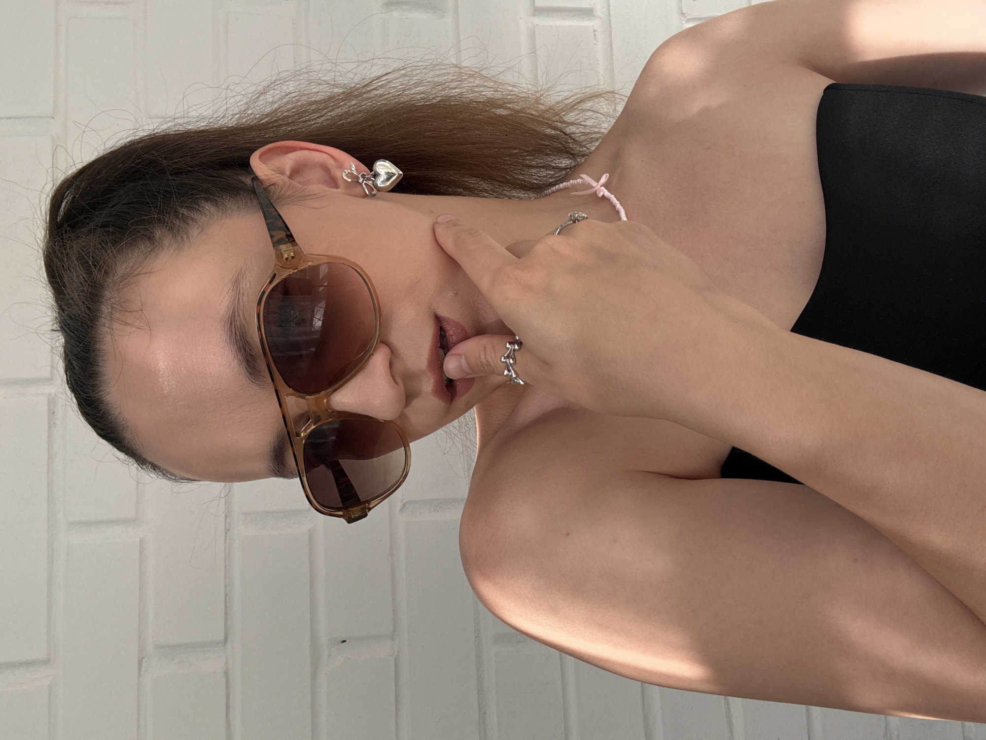 The image shows a close-up, waist-up shot of a young woman with brown hair pulled into a high ponytail. She is wearing large, tortoiseshell aviator sunglasses and has her finger to her lips. She has a silver heart-shaped earring with a bow detail in her left ear, and two silver rings on her right hand. She is wearing a strapless black top, and a thin pink necklace with a small pink bow. Her right shoulder is illuminated by a streak of sunlight. The background is a white brick wall. The image is undated and does not contain any information that would allow for its location to be specified as Chaikovsky, Russia, or any other specific city.